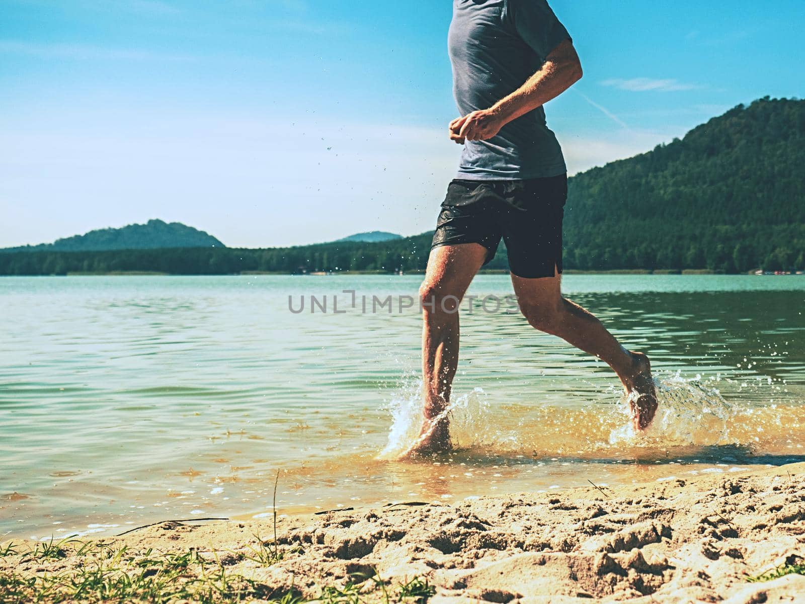 Barefoot sportsman run in water along the sandy beach. Water is splashing man legs and body.