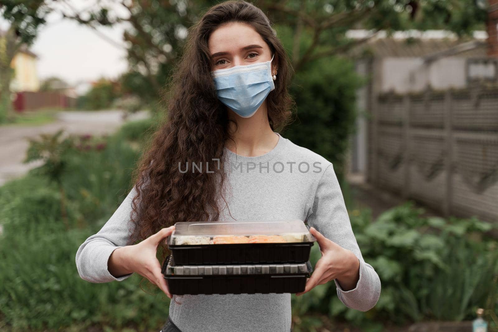 Girl courier in medical mask with 2 sushi boxes stands in front of car. Sushi set in box healthy food delivery service by car. by Rabizo