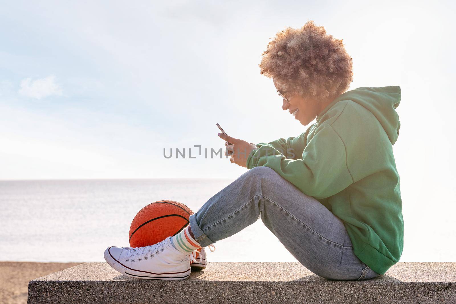 smiling young latin woman in a hoodie sitting on a bench by the beach with a basketball using her phone, concept of lifestyle and technology of communication, copy space for text