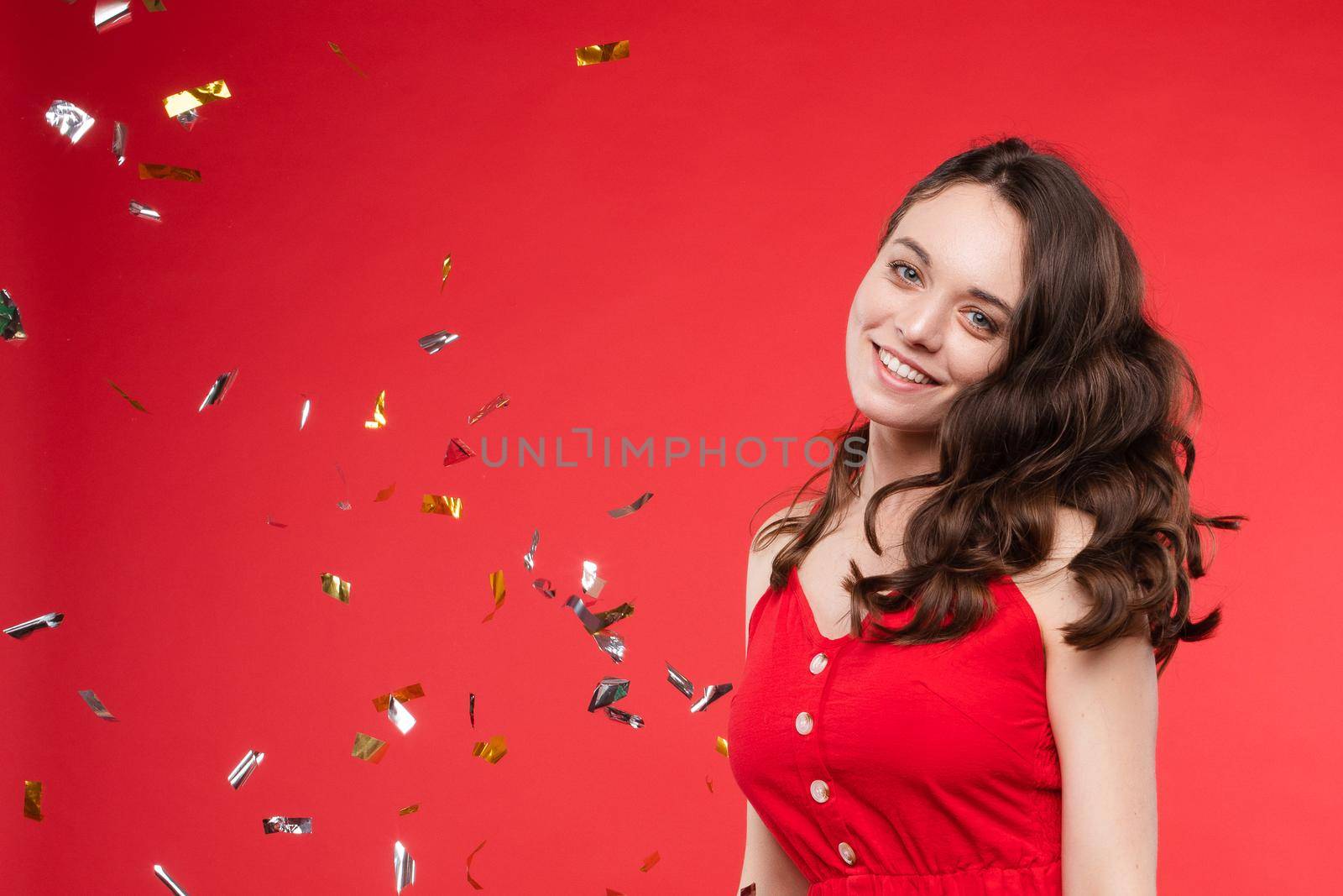 Front view of happy young girl wearing red dress looking at camera and twirling hair with hand in studio. Curly model posing and smiling on isolated background. Concept of emotions and celebration.