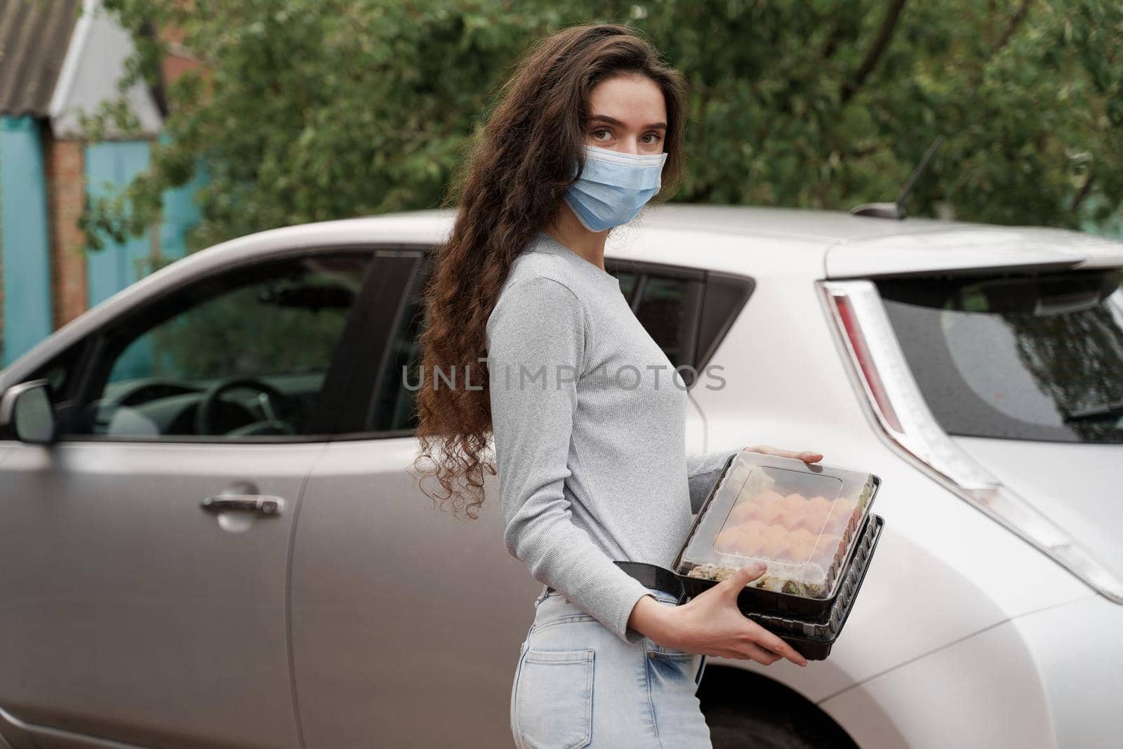 Sushi set in box healthy food delivery service by car. Girl courier in medical mask with 2 sushi boxes stands in front of car