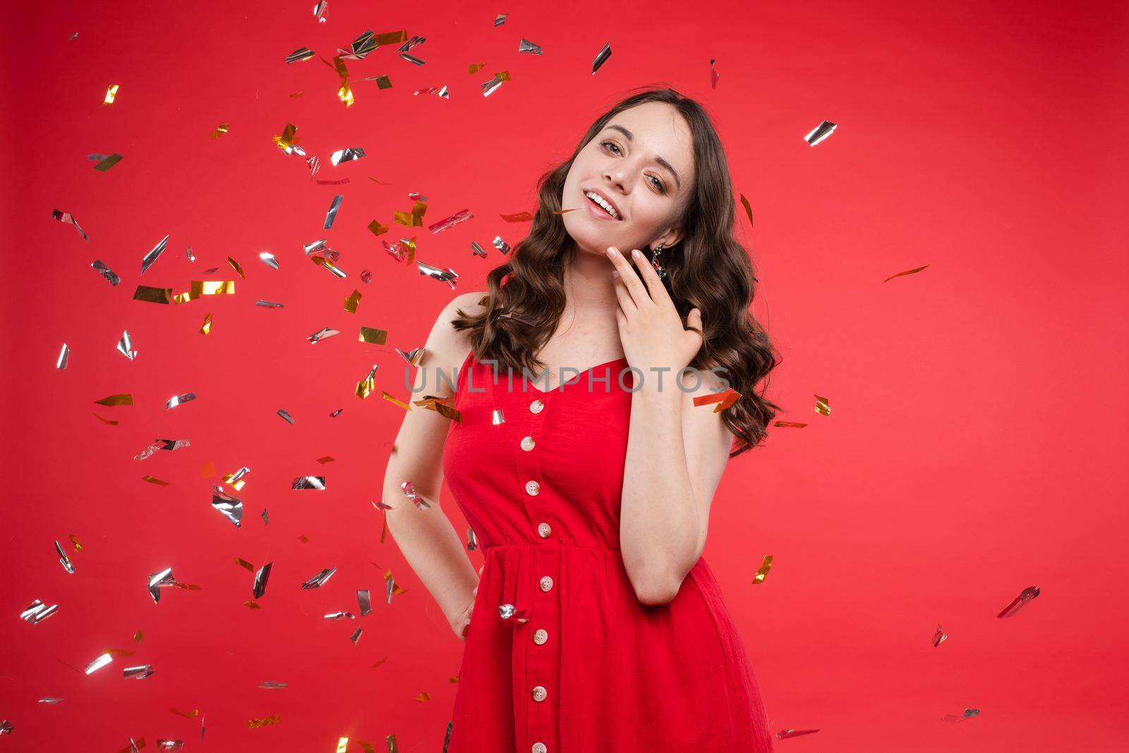 Front view of pretty brunette wearing red dress looking at camera while sparkles falling around in studio. Beautiful woman with hairstyle posing on isolated background. Concept of holiday.