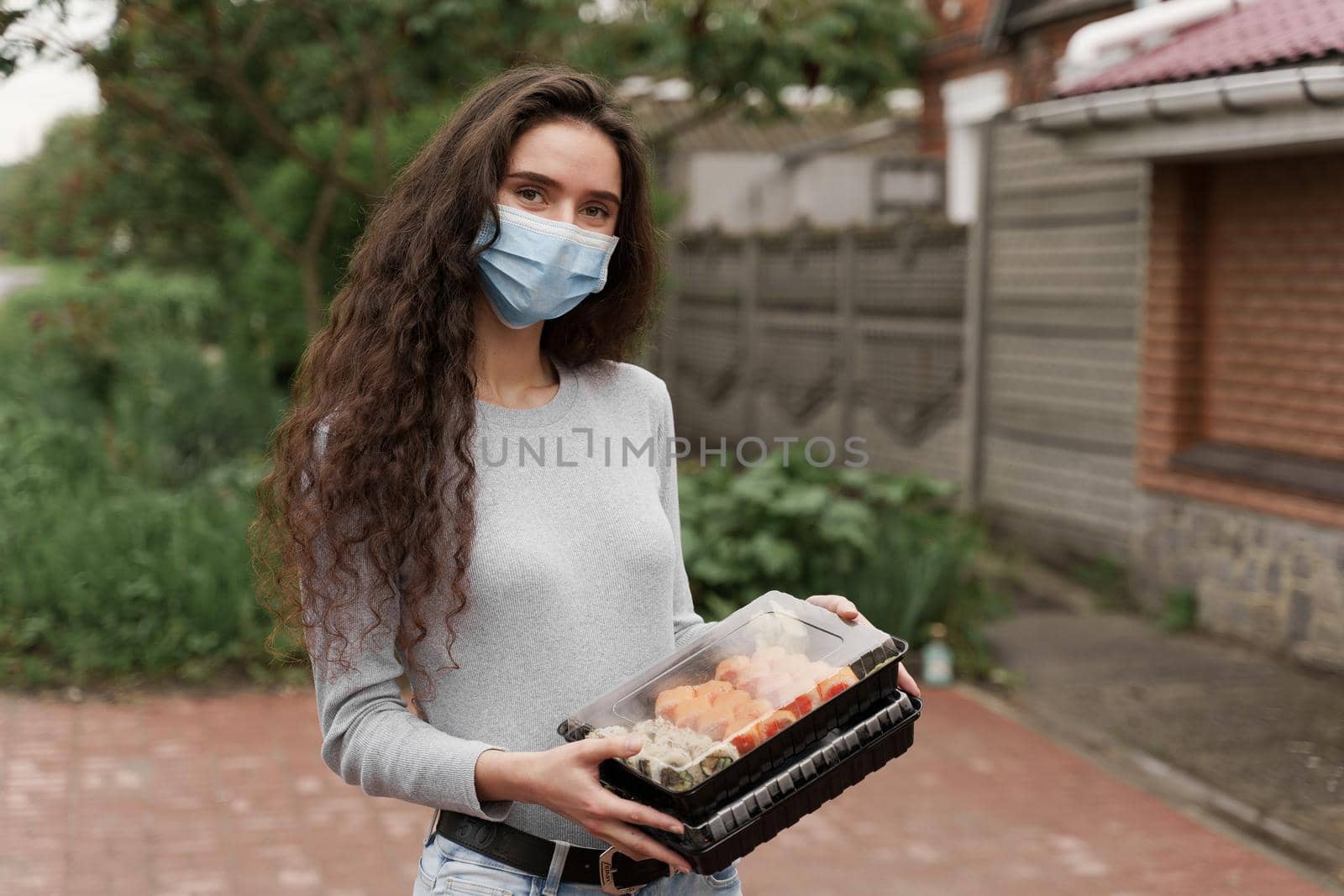 Sushi set in box healthy food delivery service by car. Girl courier in medical mask with 2 sushi boxes stands in front of car
