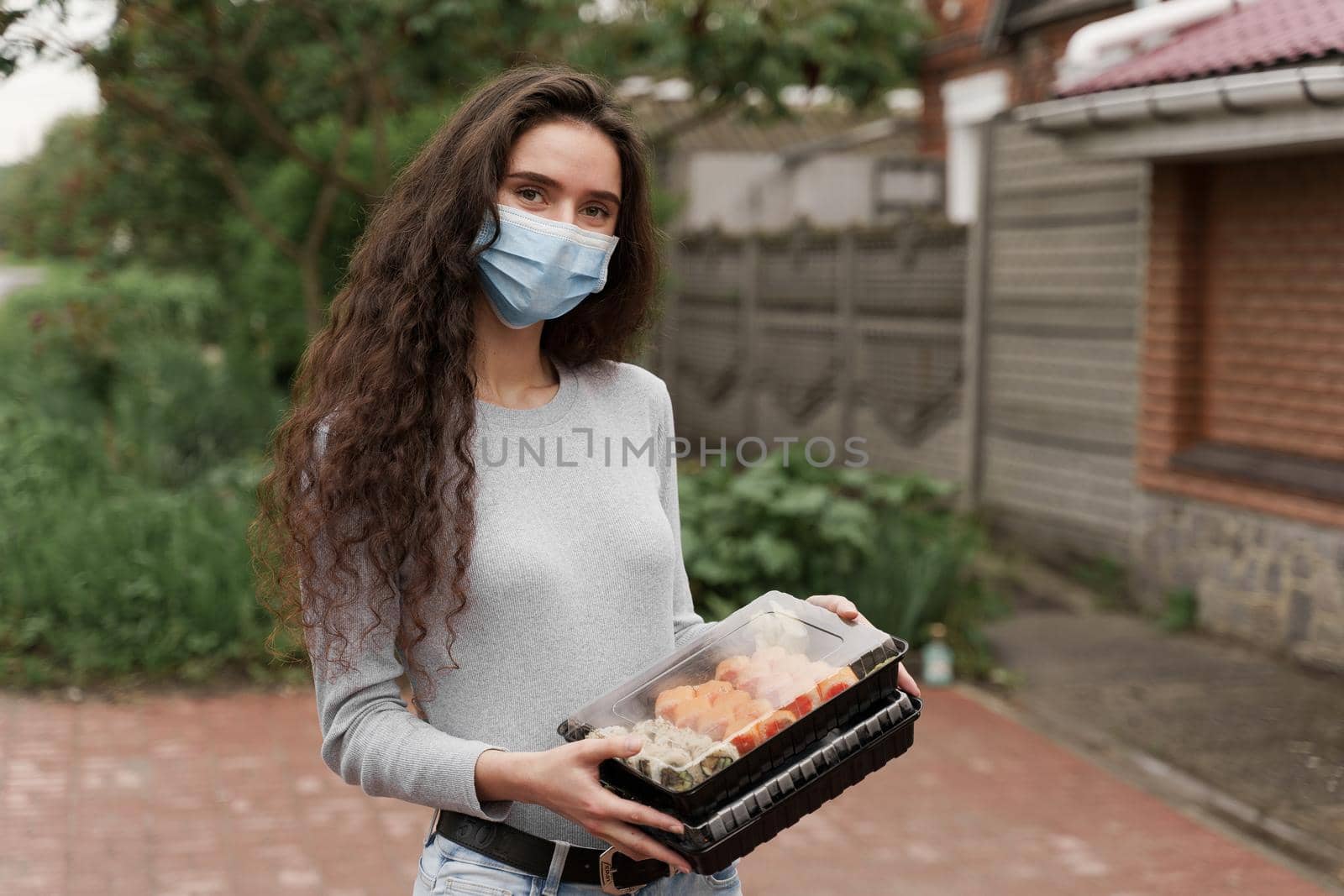 Sushi set in box healthy food delivery service by car. Girl courier in medical mask with 2 sushi boxes stands in front of car. by Rabizo