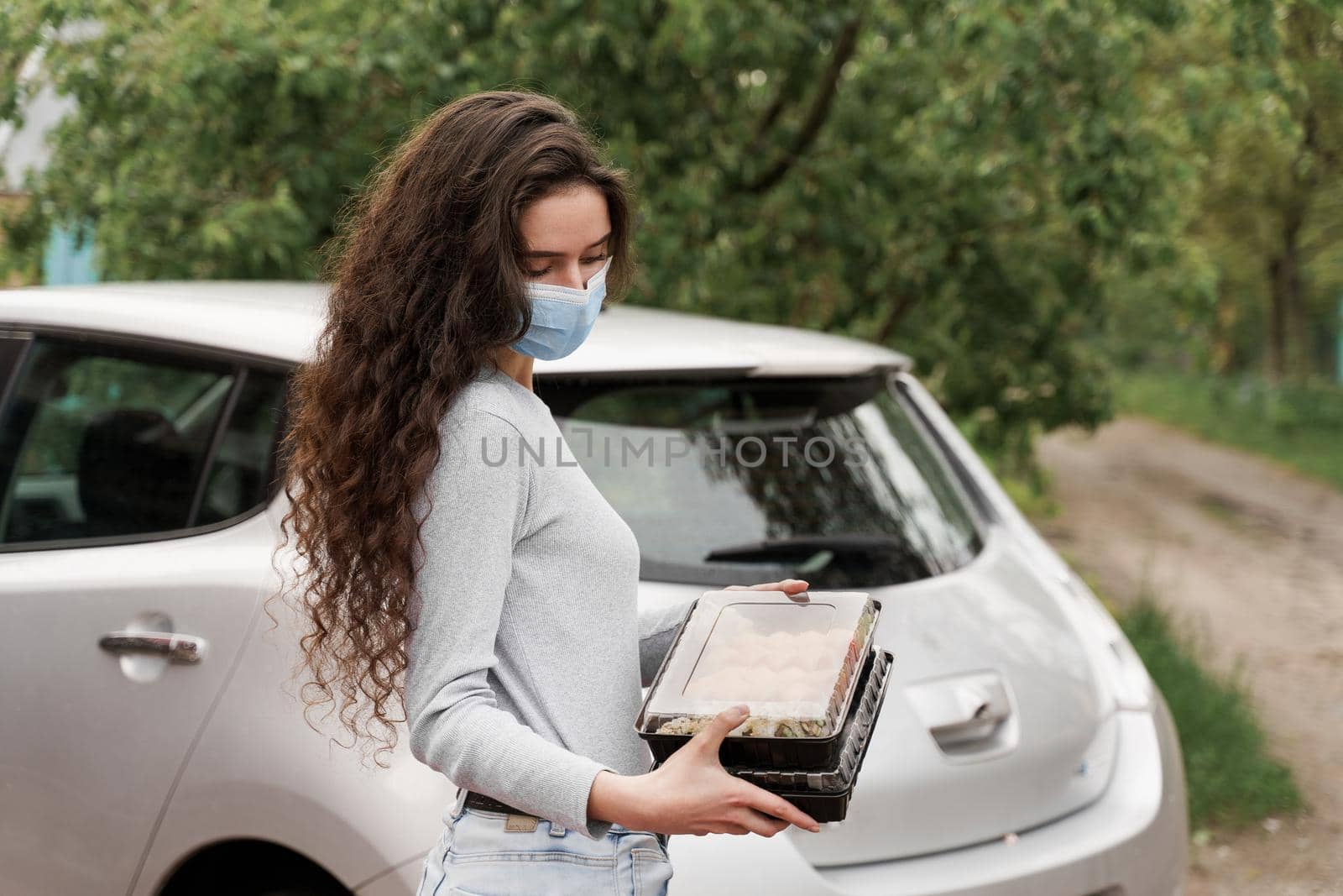 Sushi set in box healthy food delivery service by car. Girl courier in medical mask with 2 sushi boxes stands in front of car. by Rabizo