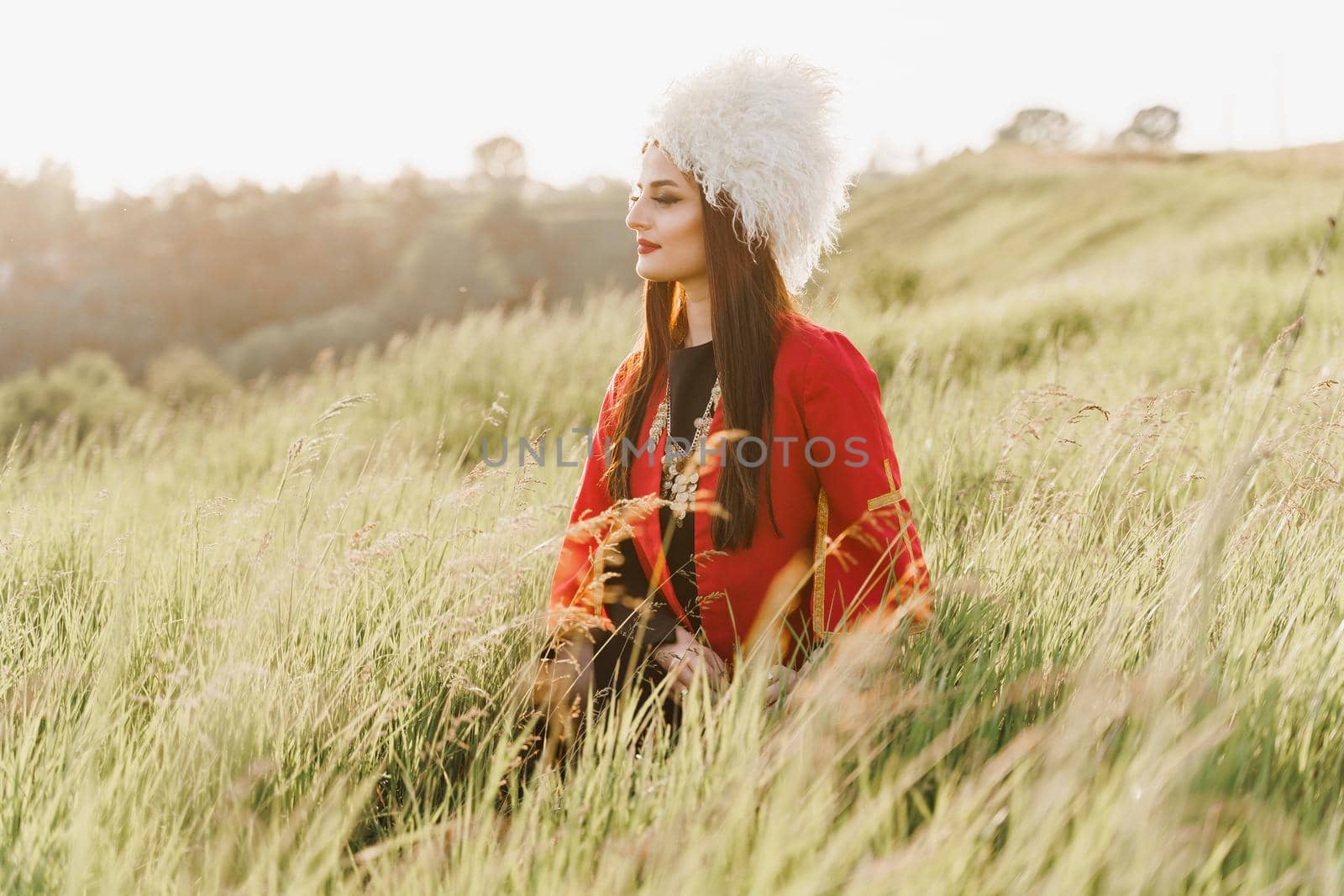 Georgian girl in white papakha and red national dress seats on the green grass and looks left side. Georgian culture lifestyle.