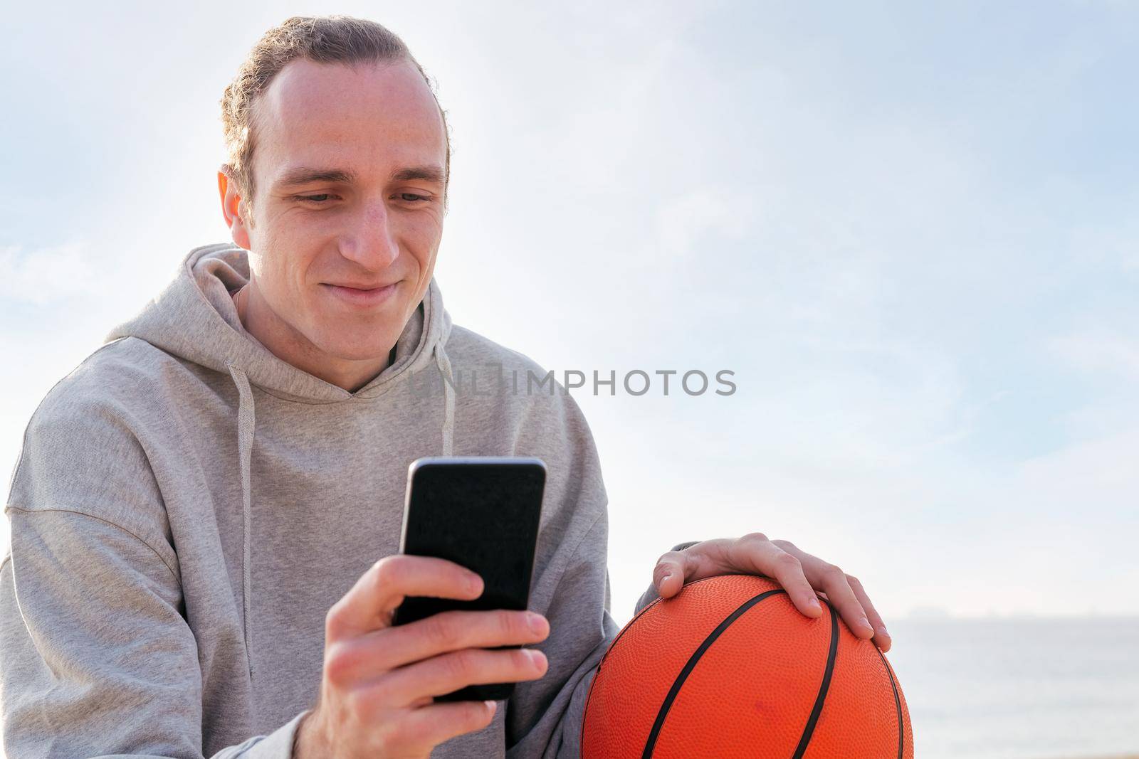 smiling caucasian man with a hoodie and a basketball using his mobile phone, concept of lifestyle and technology of communication, copy space for text