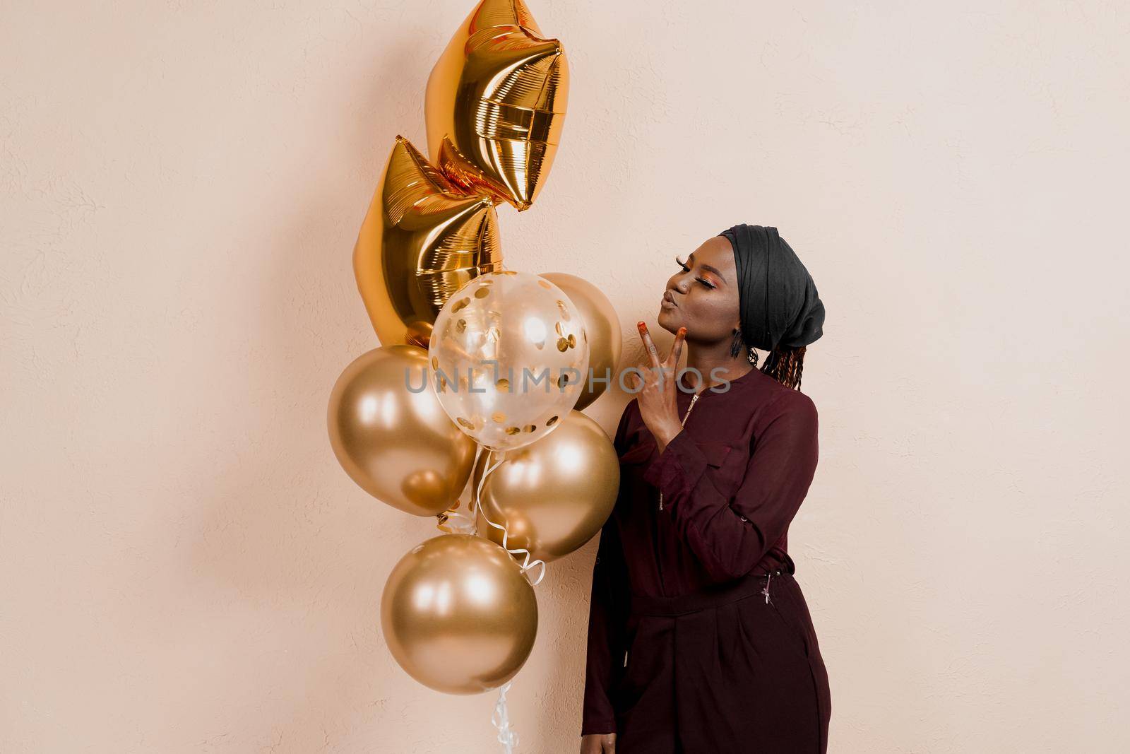 Muslim black girl with golden helium balloons isolated peach background. African young woman celebrates her birthday. by Rabizo