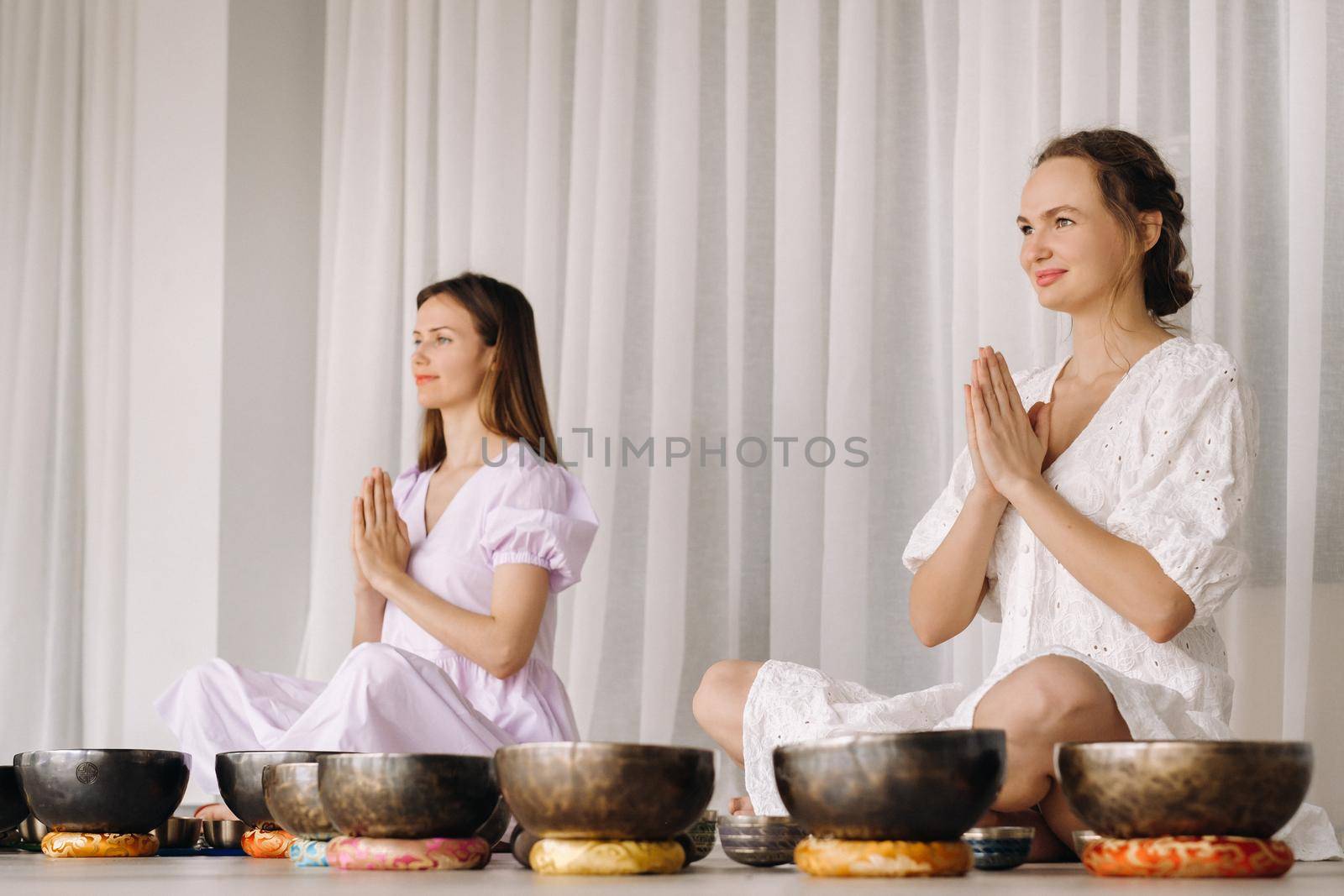 Two women are sitting with Tibetan bowls in the lotus position before a yoga class in the gym by Lobachad