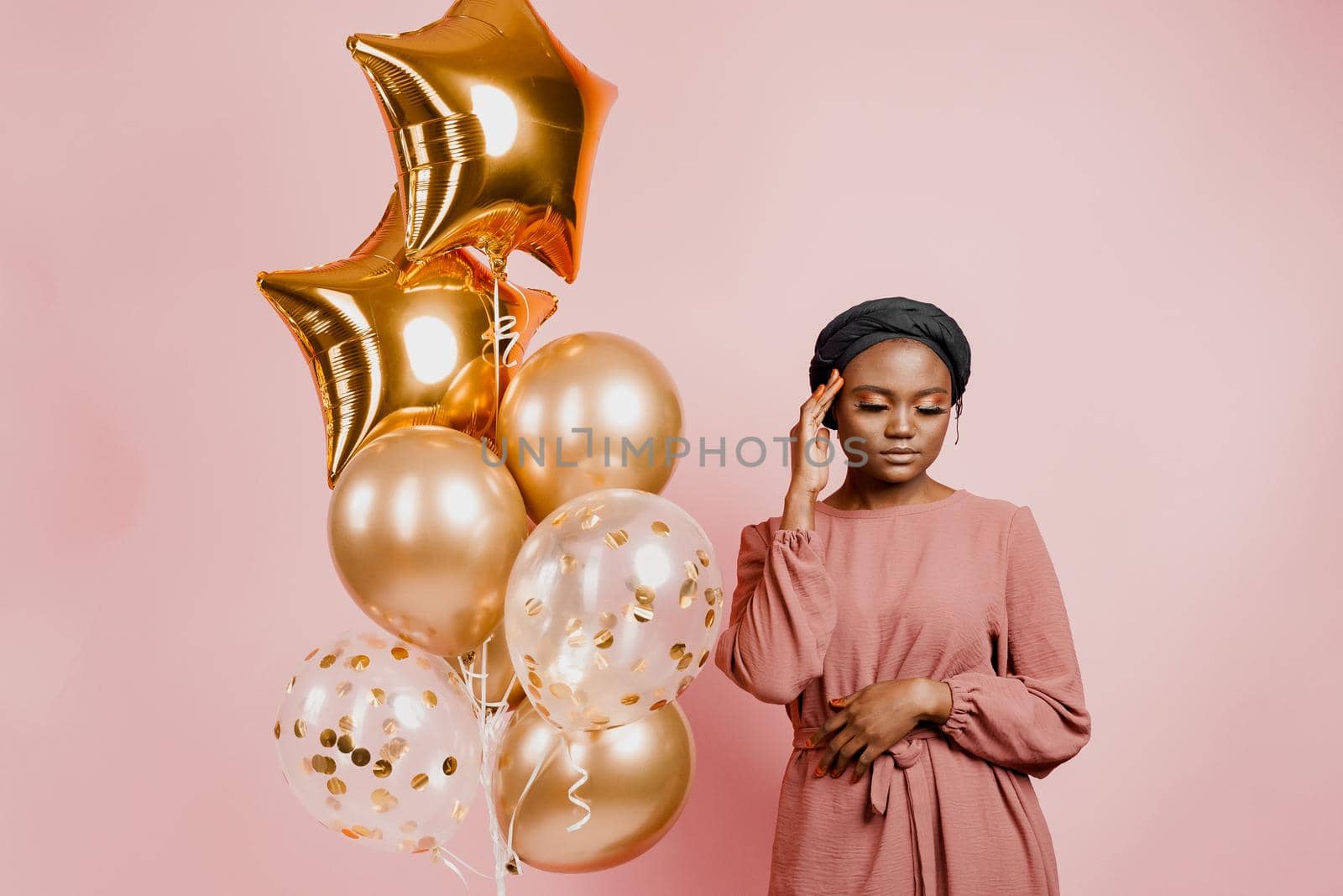 Muslim black girl with golden helium balloons smiles isolated pink background. Close-up african young woman celebrates her birthday