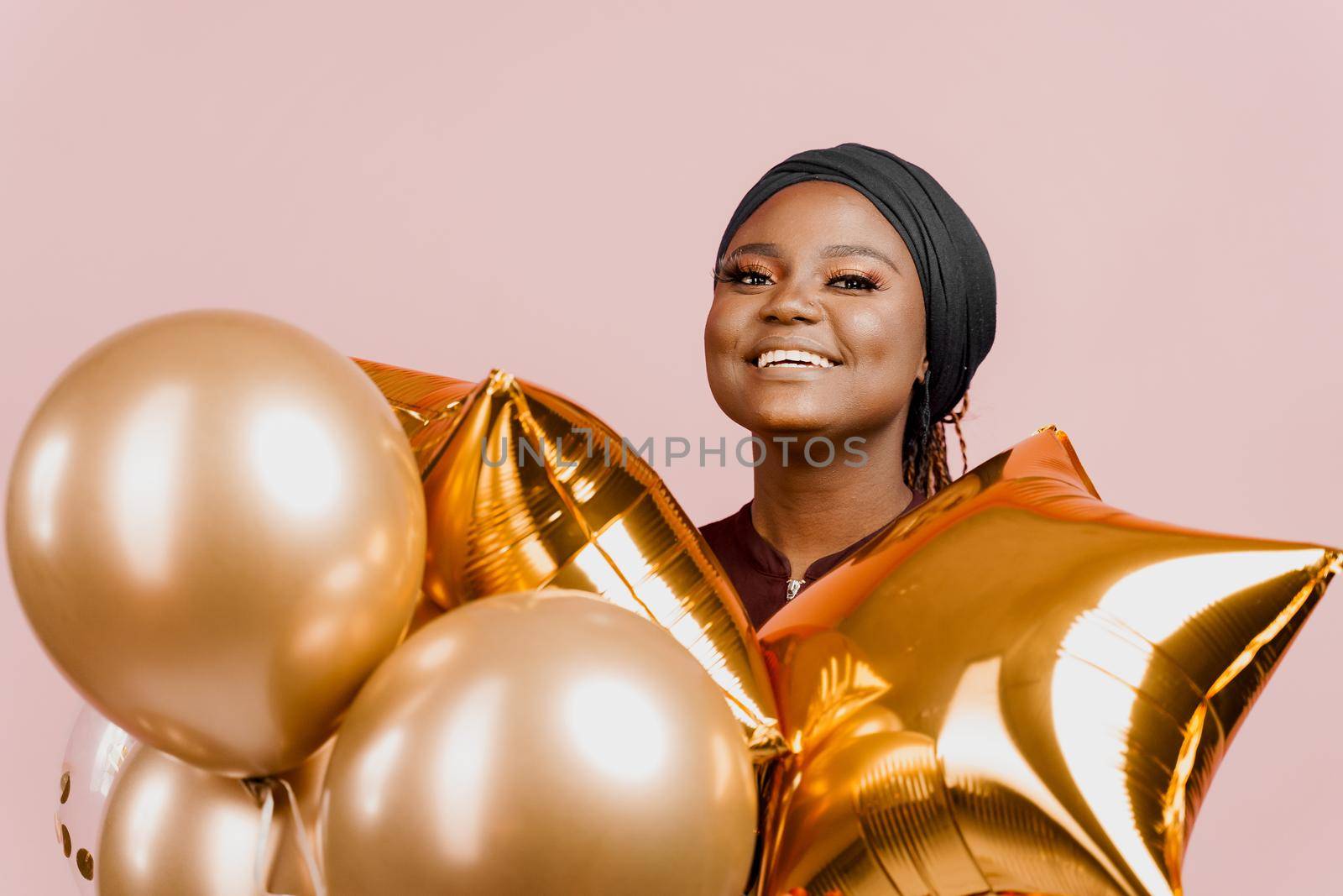 Muslim black girl with golden helium balloons smiles isolated pink background. Close-up african young woman celebrates her birthday