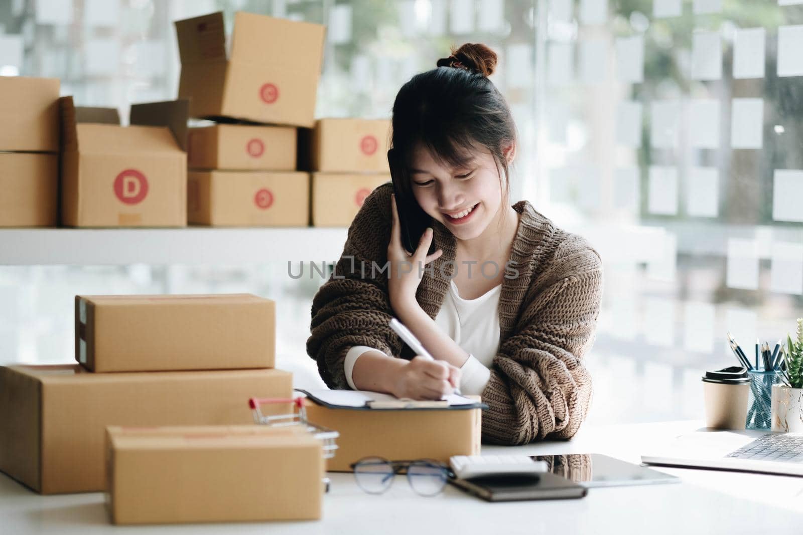 Startup small business entrepreneur SME freelance woman work with smart phone, Young Asian small business owner use computer, online market packing box delivery, SME e-commerce telemarketing concept.