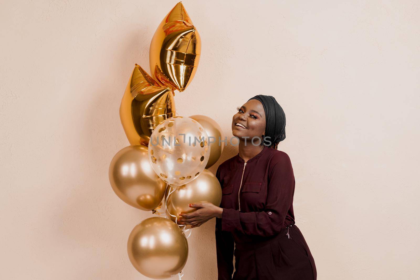 Eid al-Adha celebration. Muslim national holiday. Attractive girl with golden helium balloons isolated peach background. Young black woman weared traditional dress and scarf by Rabizo
