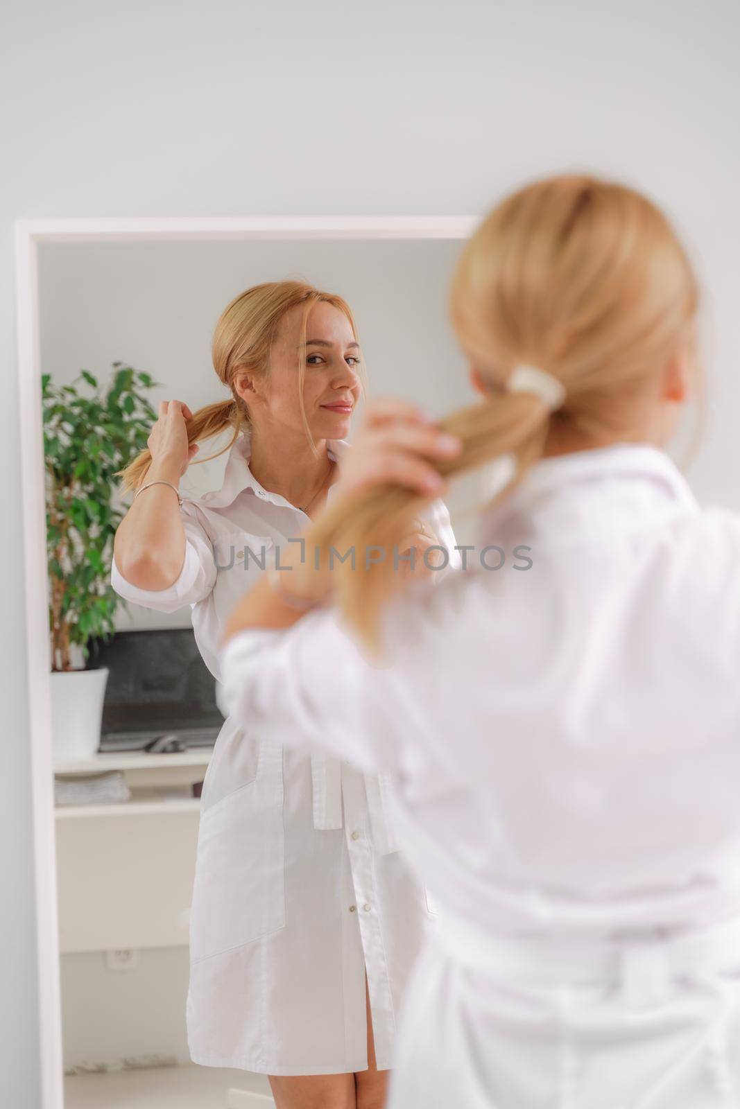 A blonde woman in white formal clothes looks at herself in the mirror. On a white background, green leaves at the back. by Matiunina