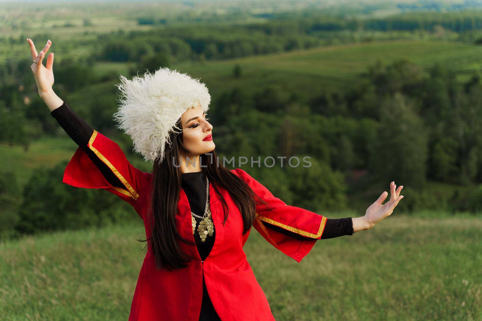 Georgian girl in white papakha dances national dance in red national dress on the green hills of Georgia background. Georgian culture lifestyle.