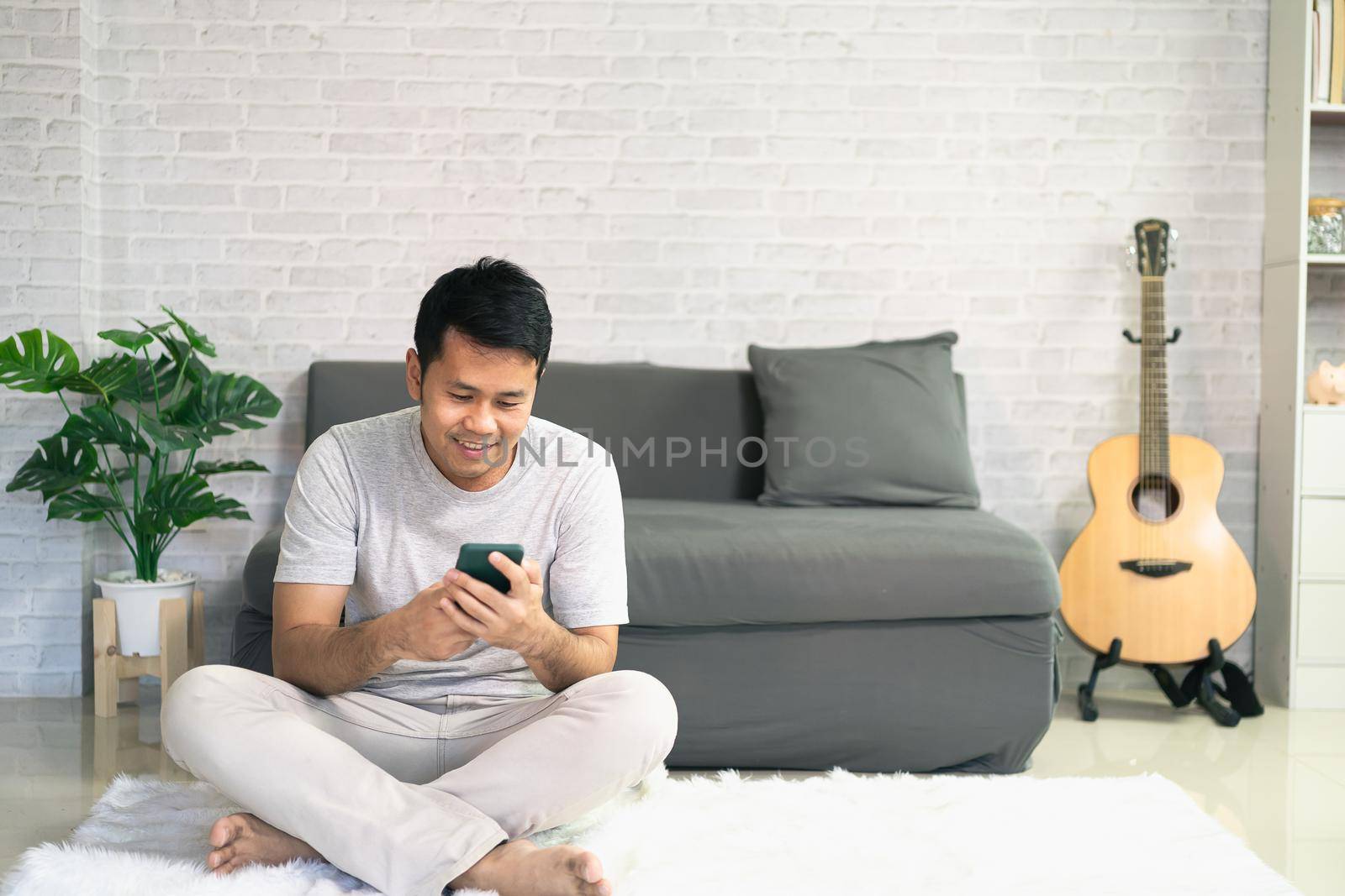 Smiling asian man wearing casual clothes sitting on a couch at the living room. Asian man using mobile phone chatting in living room. by Wmpix