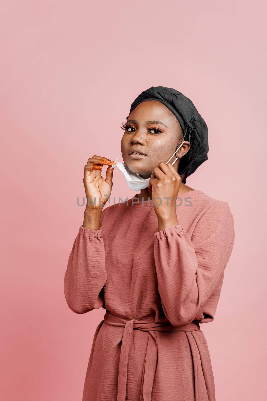 Muslim girl in medical mask on the pink background. Portrait of african woman who sick coronavirus covid-19. Black girl touches her mask. Vertical photo. by Rabizo