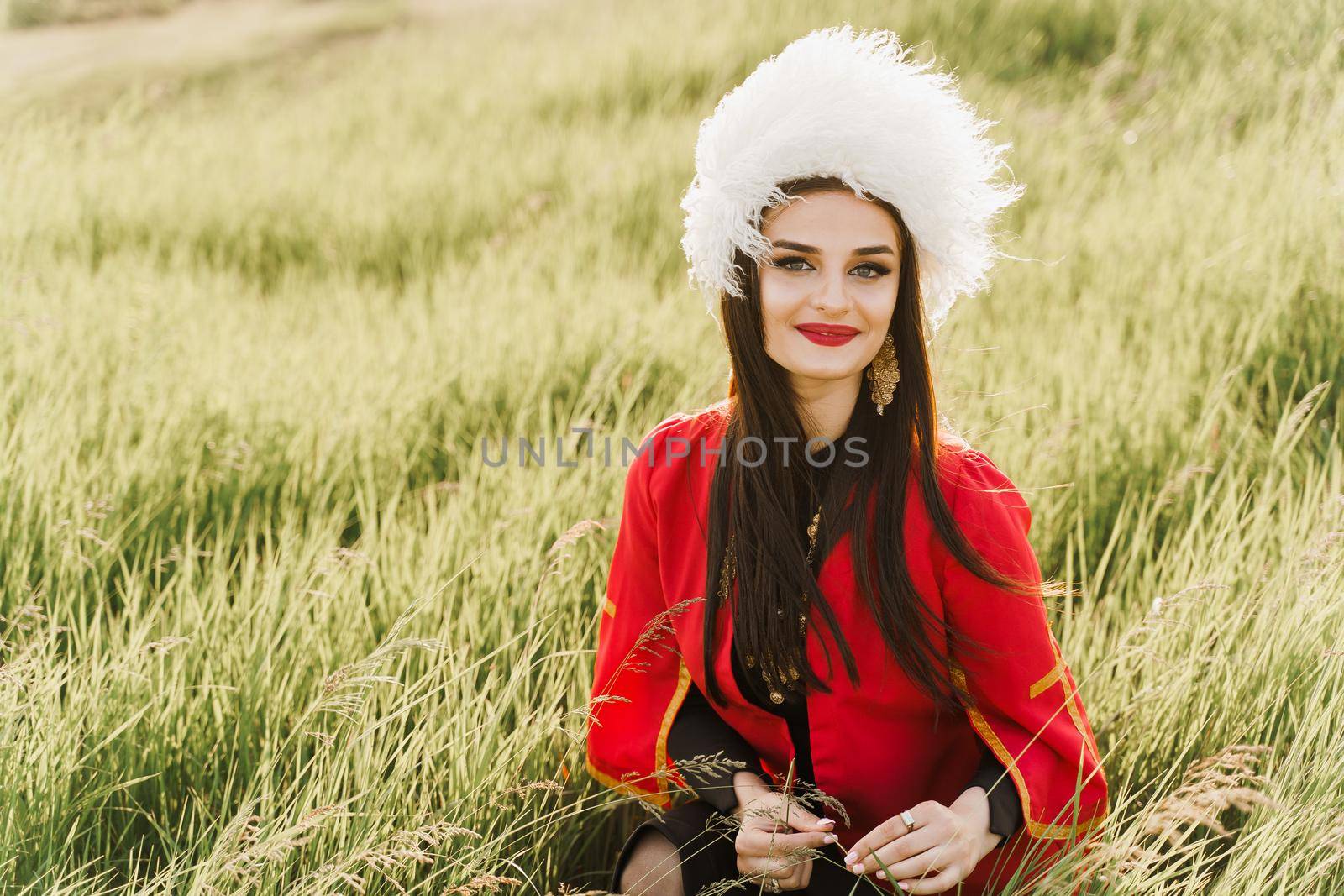 Georgian girl in white papakha and red national dress seats on the green grass and smiles into camera. Georgian culture lifestyle by Rabizo