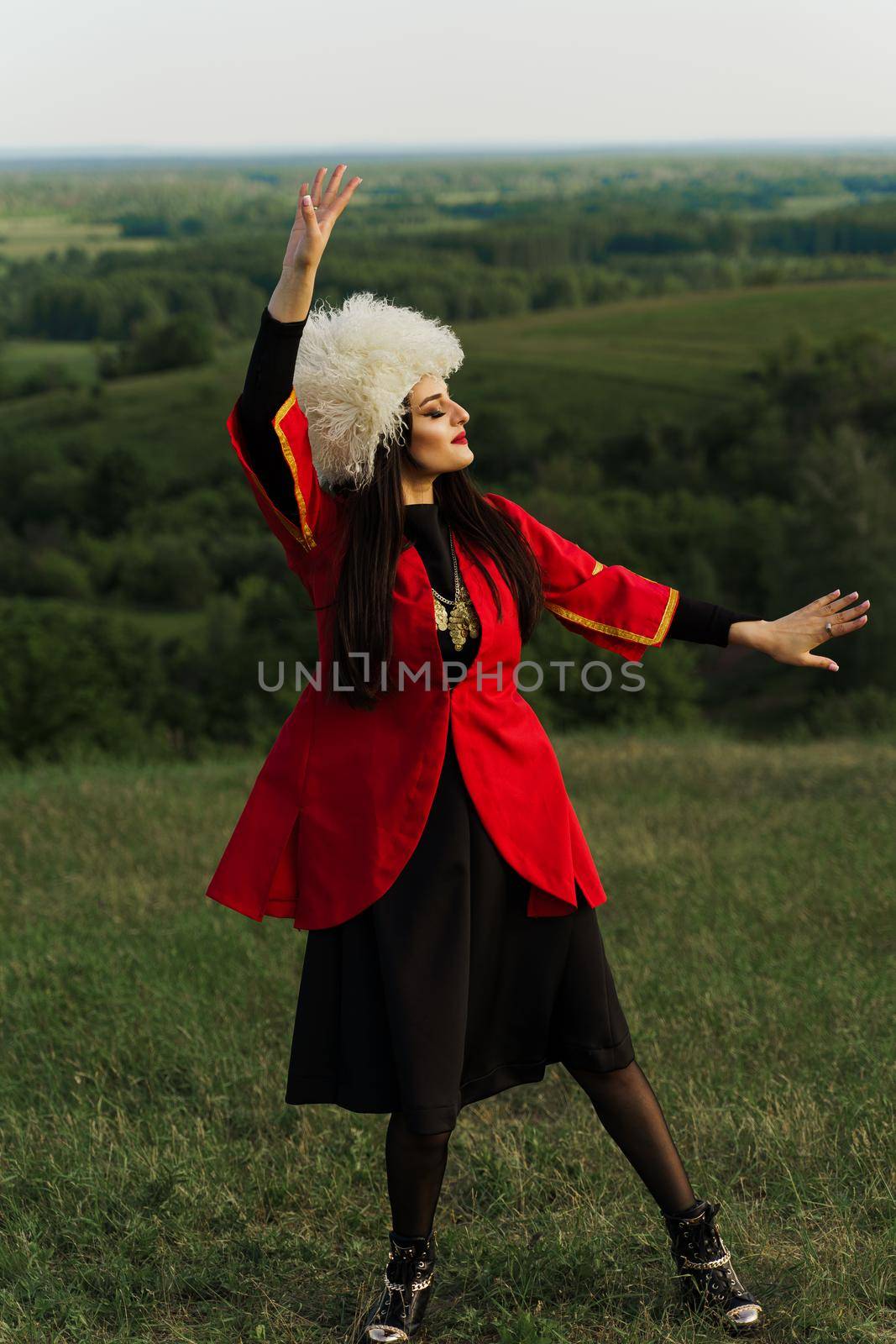 Georgian girl in white papakha and red national dress dances national dance named: rachuli, acharuli, osuri, shalaxo on the green hills of Georgia