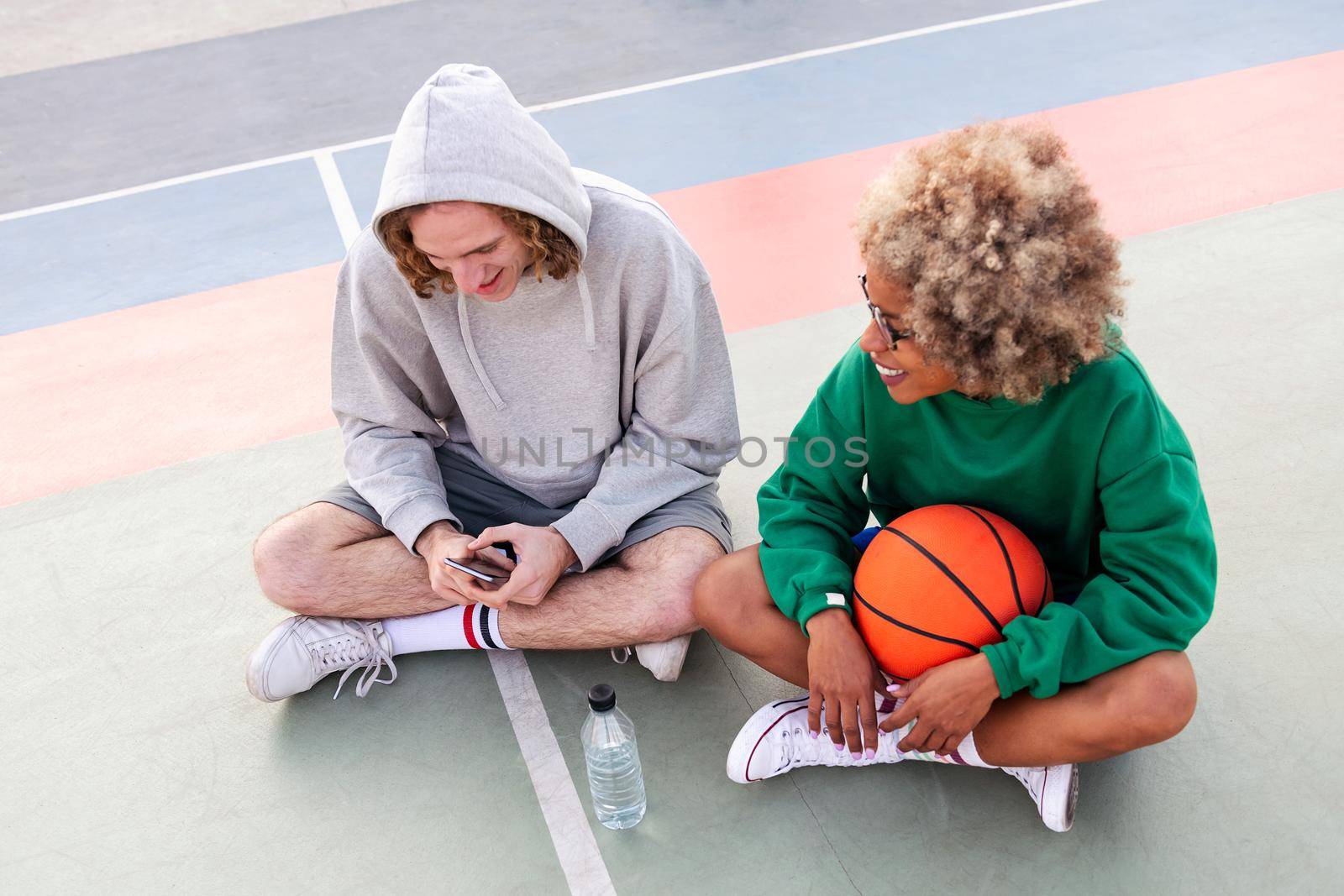 couple of friends chatting and sharing a good time sitting on the court after a basketball practice at a city park, concept of friendship and urban sport in the street