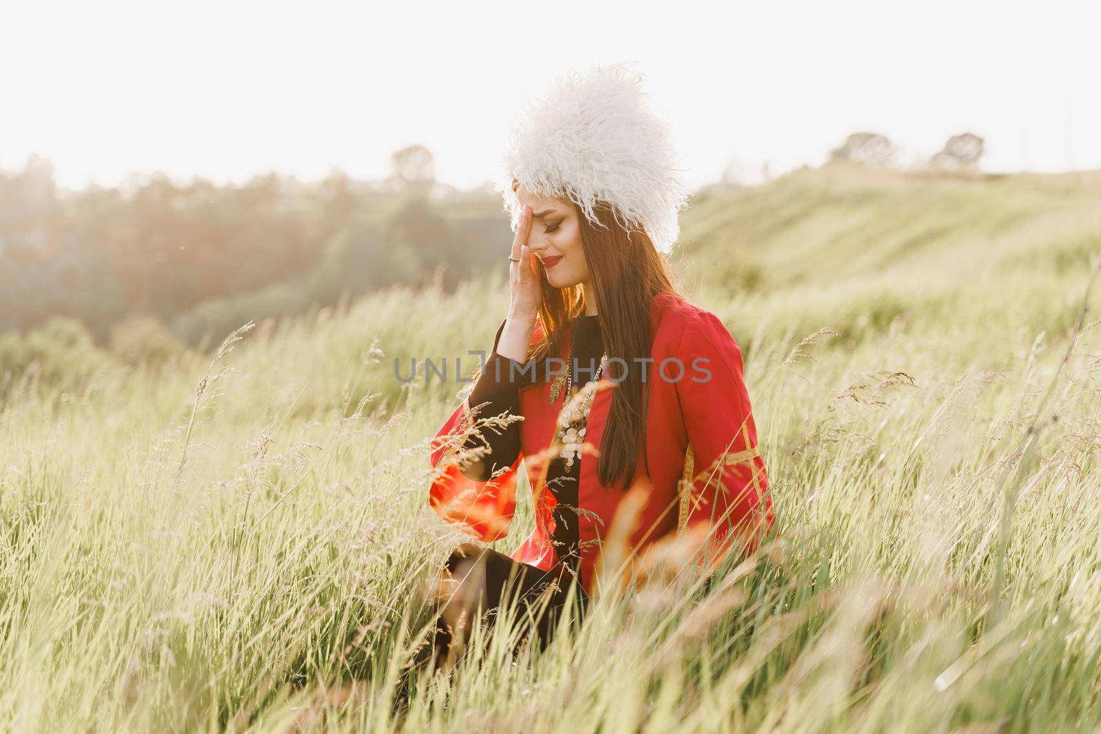 Georgian girl cry in white papakha and red national dress seats on the green grass and looks left side. Georgian culture lifestyle.