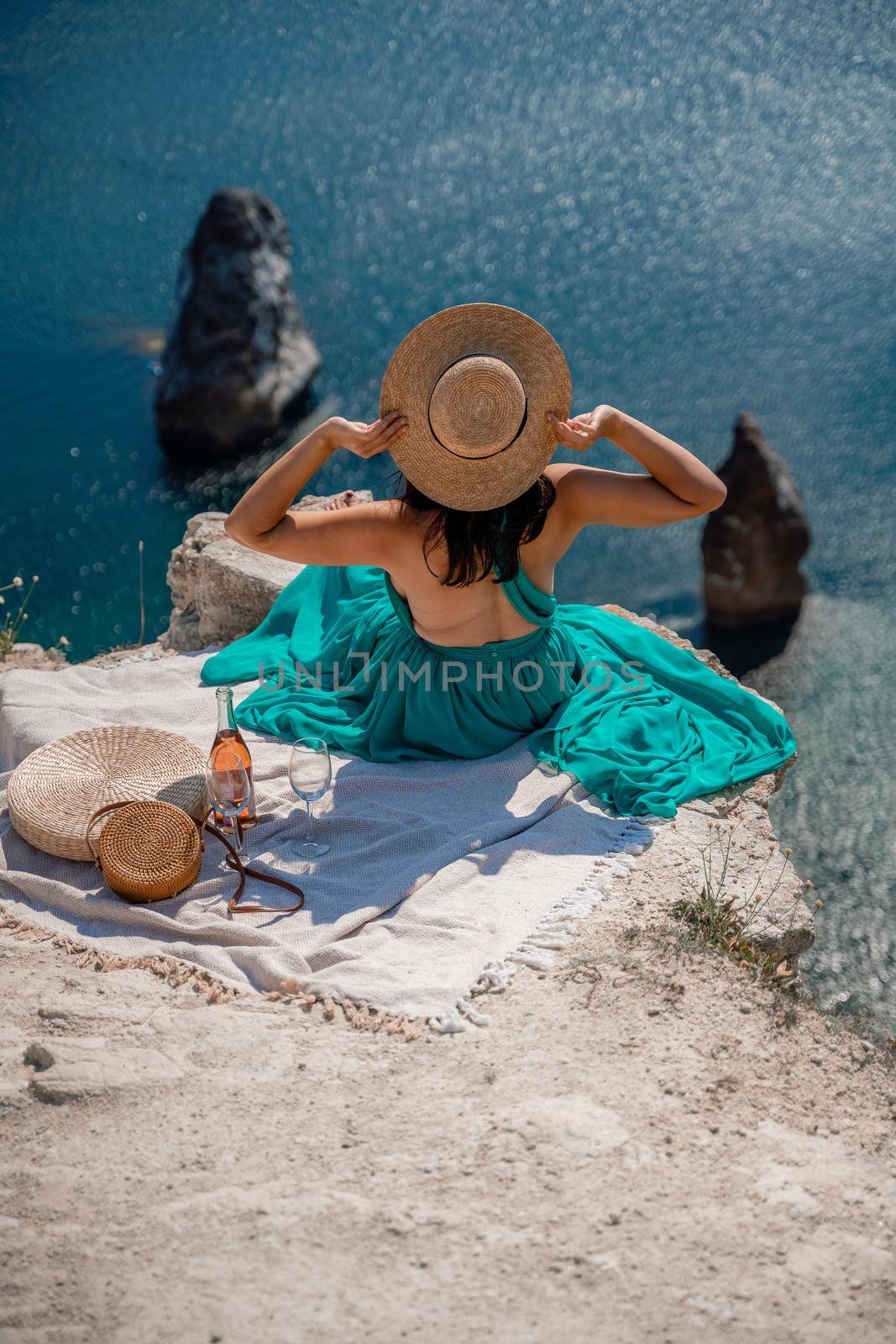 A girl in a hat sits with her back to the viewer on a picnic blanket in a mint dress and holds a hat with her hands. Champagne, two glasses, Summit on the mountain against the background of the sea and rocks in the sea. by Matiunina