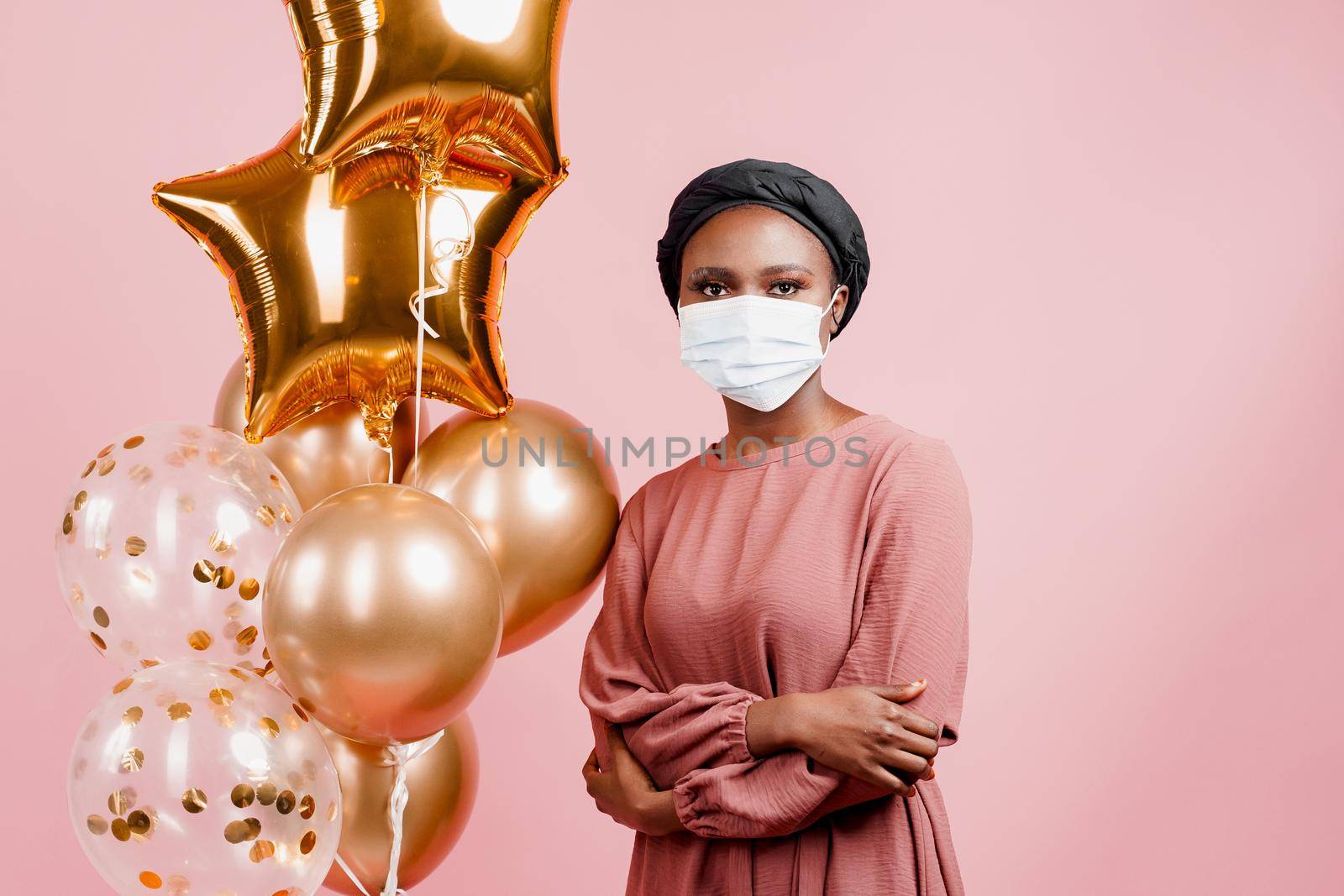 Muslim girl in medical mask on the pink background with golden baloons. Portrait close-up of african woman who sick coronavirus covid-19