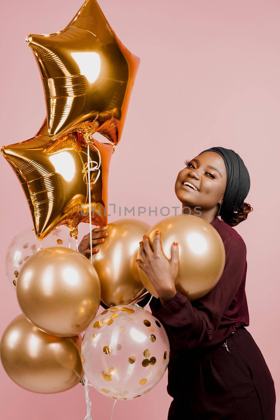 Black girl with golden ballons have a party and smile isolated on pink background. African woman celebrate graduation. Happy emotions of muslim young woman. by Rabizo
