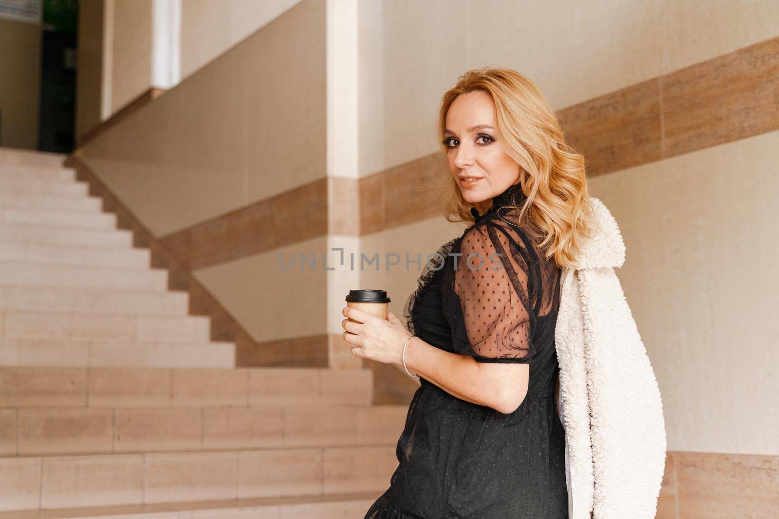 Portrait of a beautiful smiling blonde. She's walking up the stairs, coffee in hand. The Concept Of Urban Life. by Matiunina