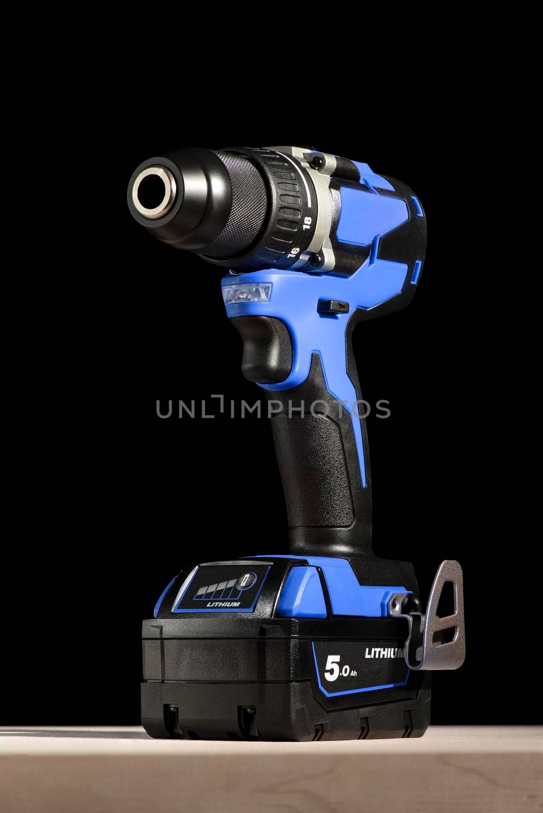 Cordless drill stands on a wooden table on a black background. Cordless drill with lithium-ion battery in blue. Professional tool for drilling holes and driving screws