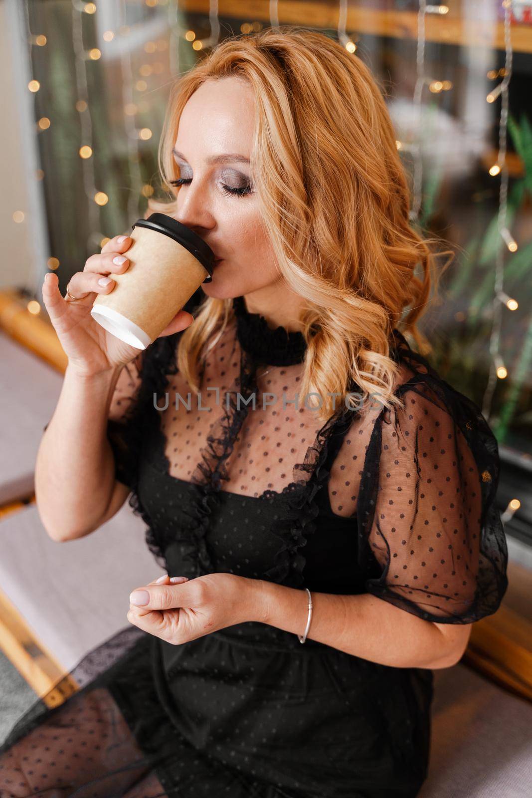 Young charming blonde with a cute smile and makeup while relaxing in a cafe. She is holding a cup of coffee in her hands. She is dressed in a black dress with transparent sleeves. by Matiunina