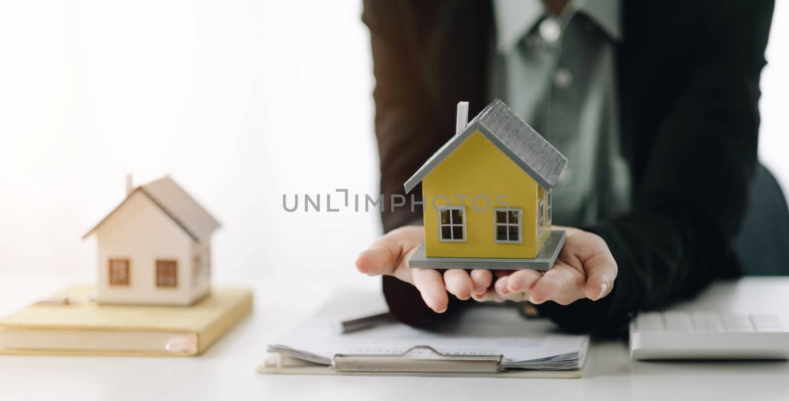 Miniature house in the hands of an Asian woman real estate agent home loan working at the office. Looking at the camera. by wichayada