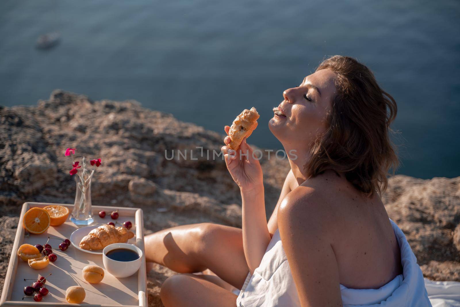 Woman covered with a blanket of bed relaxing and watching the seascape at sunrise. She holds a cup of coffee in her hand in front of her is a table with fruits and croissants. Wanderlust and freedom concept scene. by Matiunina