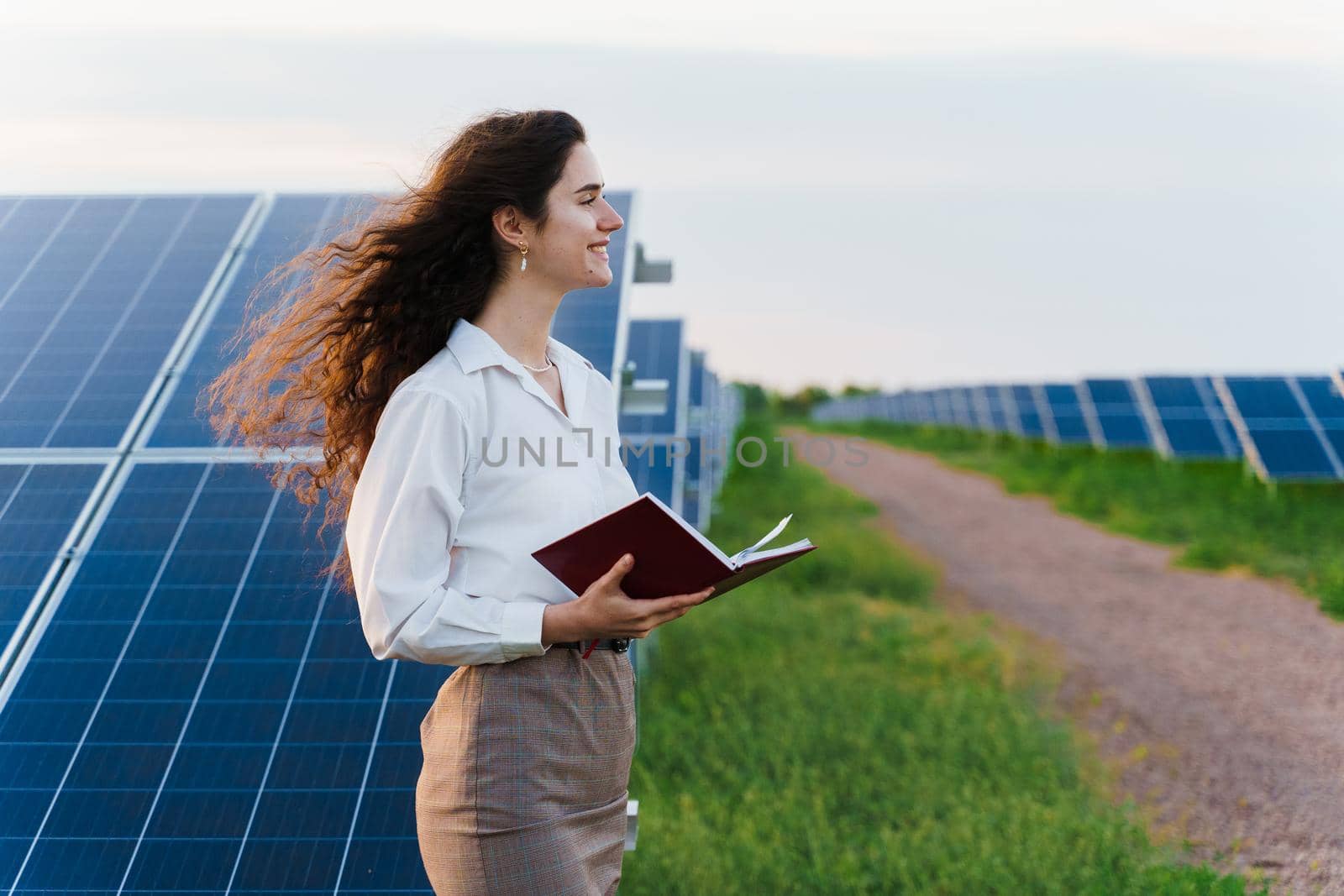 Model with solar panels stands in row on the ground. Girl dressed white formal shirt smiles on the power plant. by Rabizo