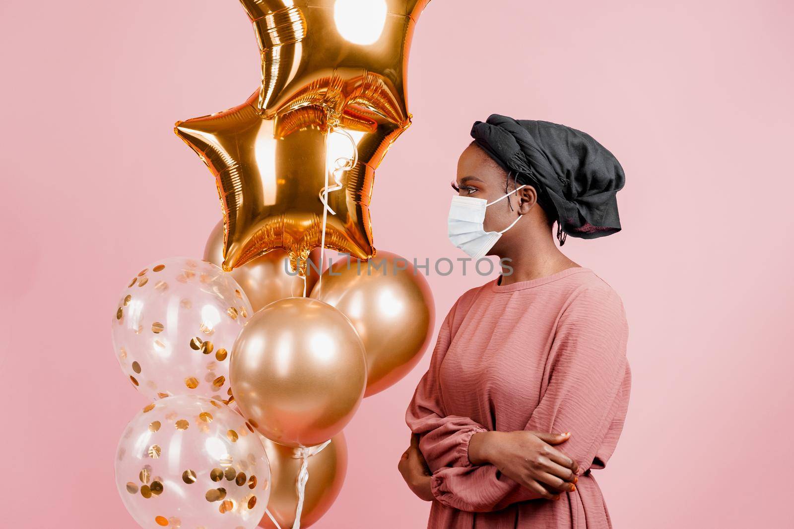 Muslim girl in medical mask on the pink background with golden baloons. Portrait close-up of african woman who sick coronavirus covid-19. by Rabizo