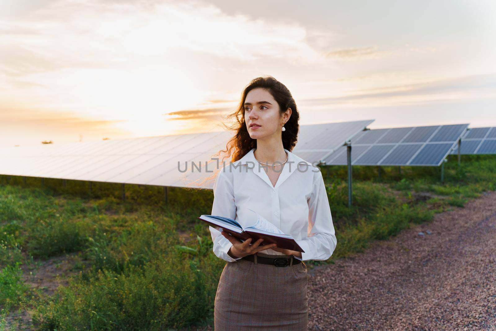 Investor woman stand and read book near solar panels row on the ground at sunset. Sustainability of planet. by Rabizo