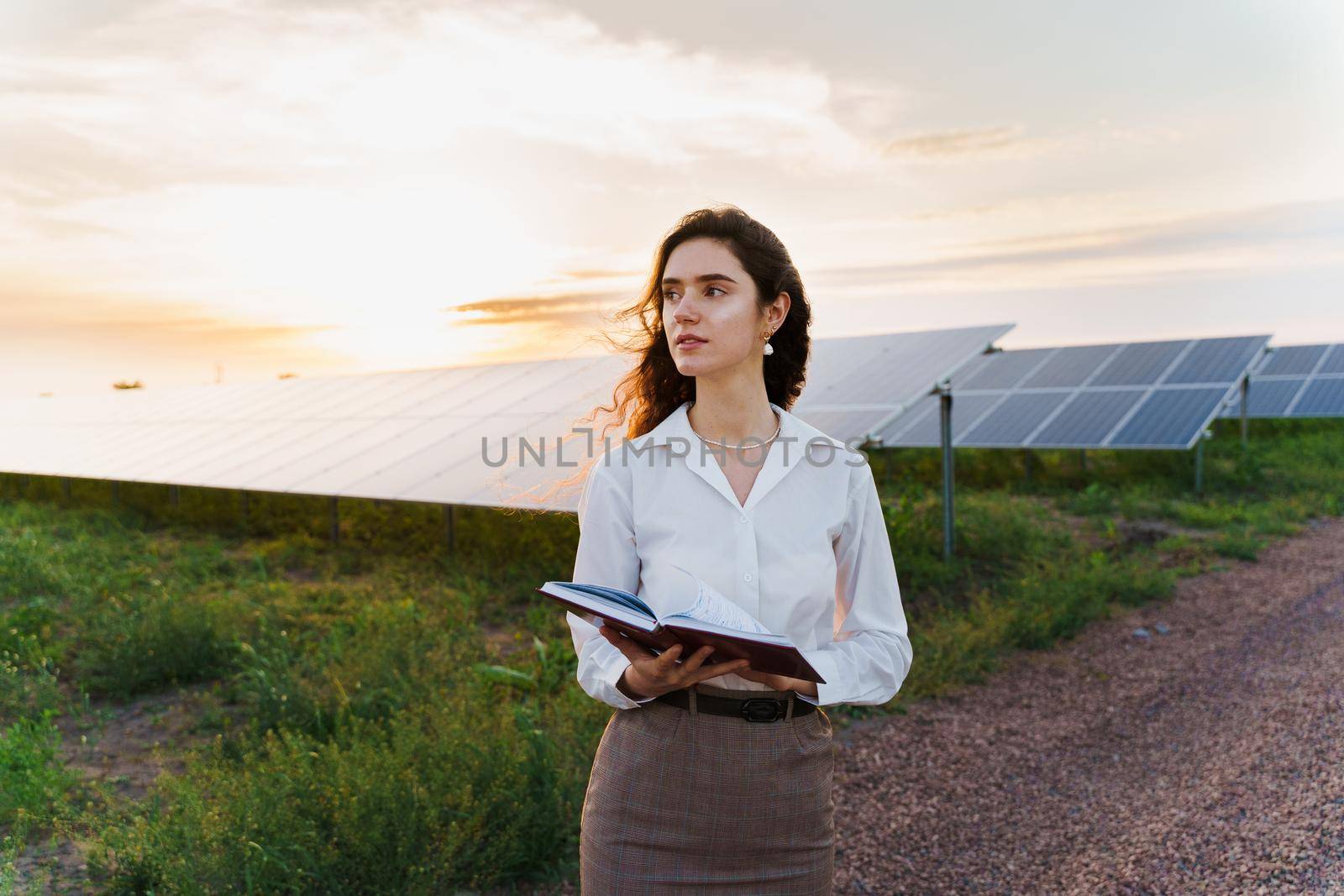 Investor woman stand and read book near solar panels row on the ground at sunset. Sustainability of planet