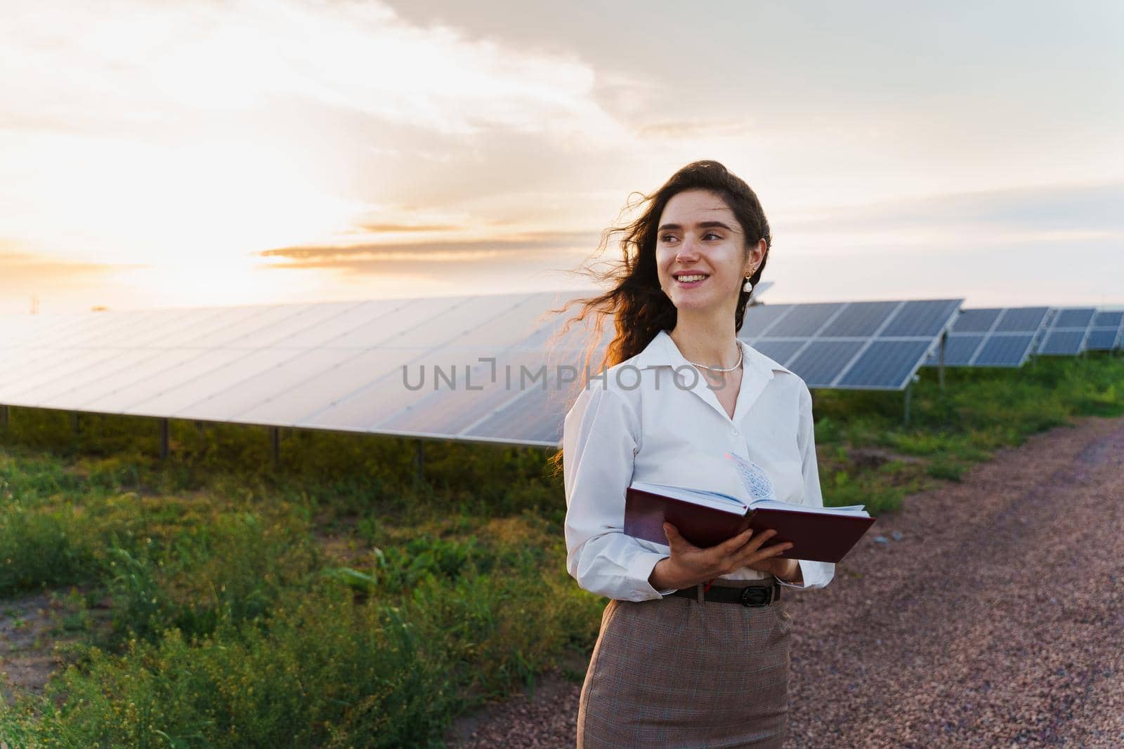 Investor woman stand and read book near solar panels row on the ground at sunset. Sustainability of planet. by Rabizo