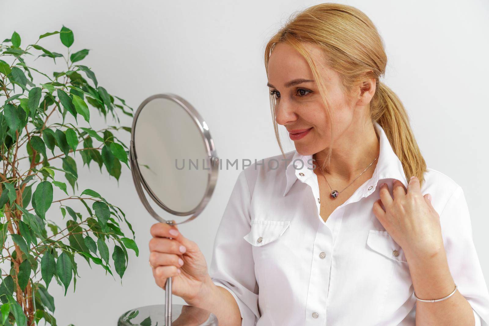 A blonde woman in white formal clothes looks at herself in the mirror. On a white background, green leaves at the back
