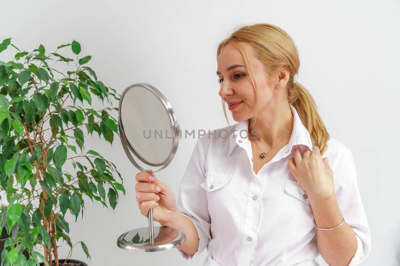 A blonde woman in white formal clothes looks at herself in the mirror. On a white background, green leaves at the back