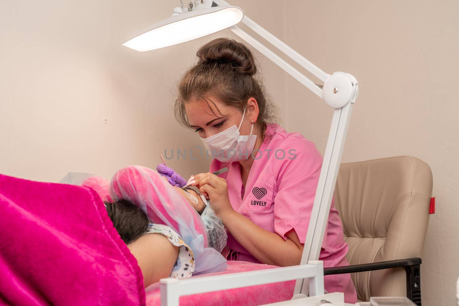 Master lashmaker, diligently extends eyelashes to the client, on the couch, in the beauty salon. A beauty master, a girl of 30 years old, European appearance, at work, in a bright room.