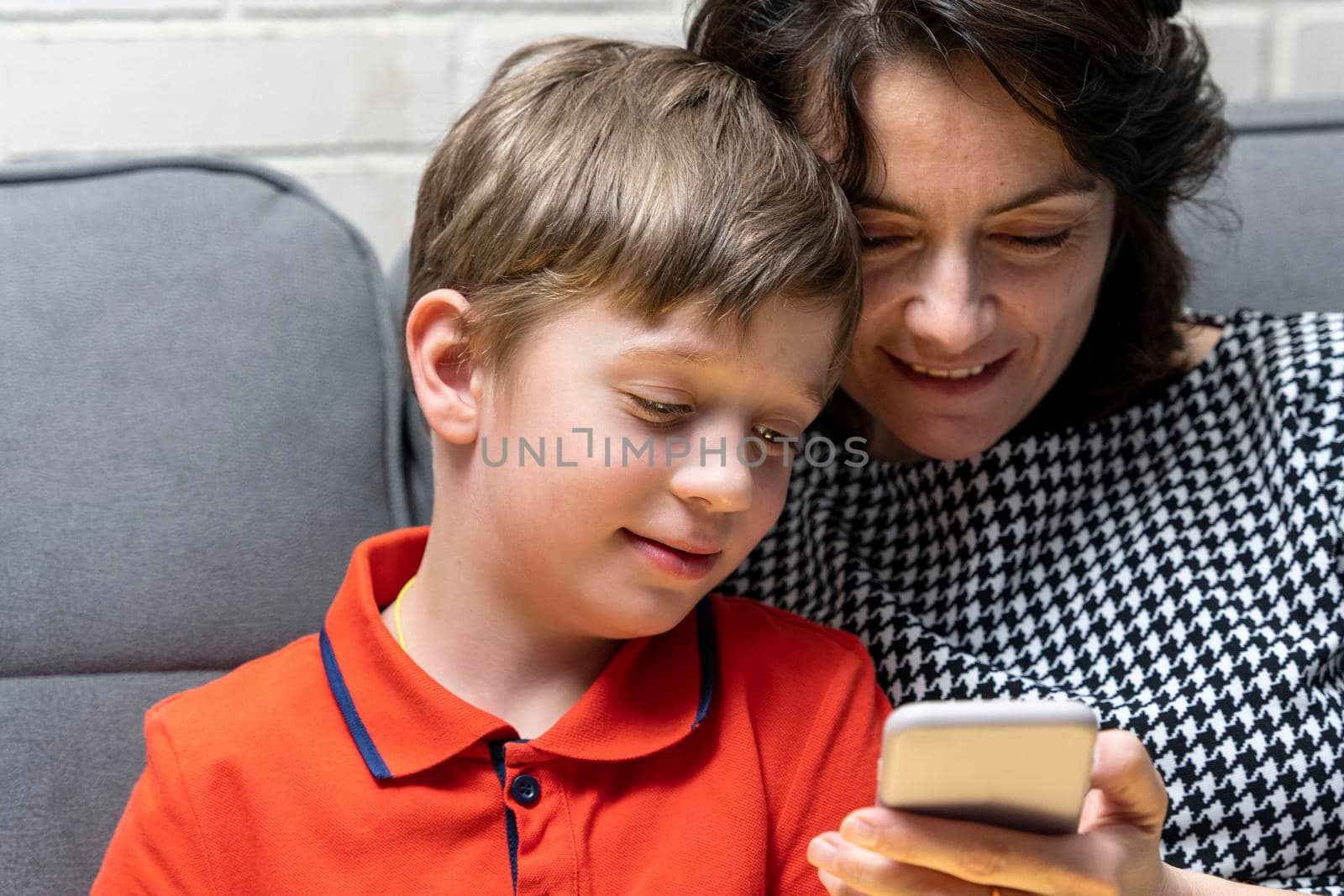 happy European preschooler boy and his mom are sitting on the couch and looking at the smartphone screen, reading congratulations, chatting online. Dependence on gadgets