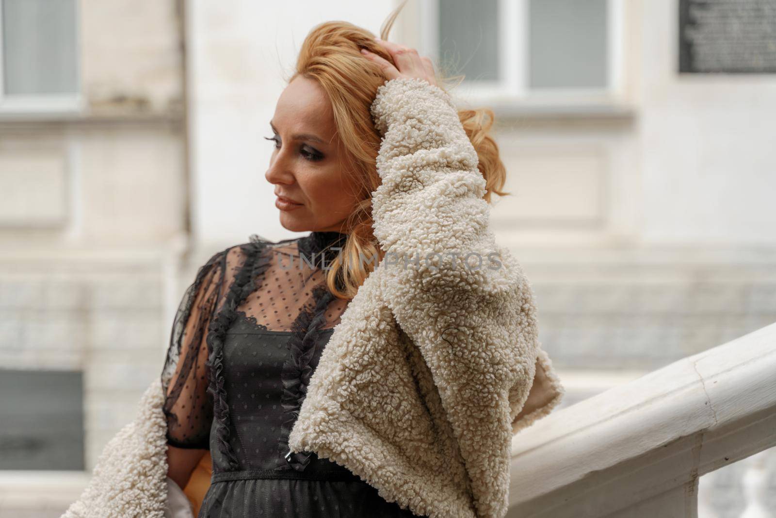 Profile of a beautiful blonde. She looks away and smiles. She is dressed in a black lace dress and a milky faux fur coat