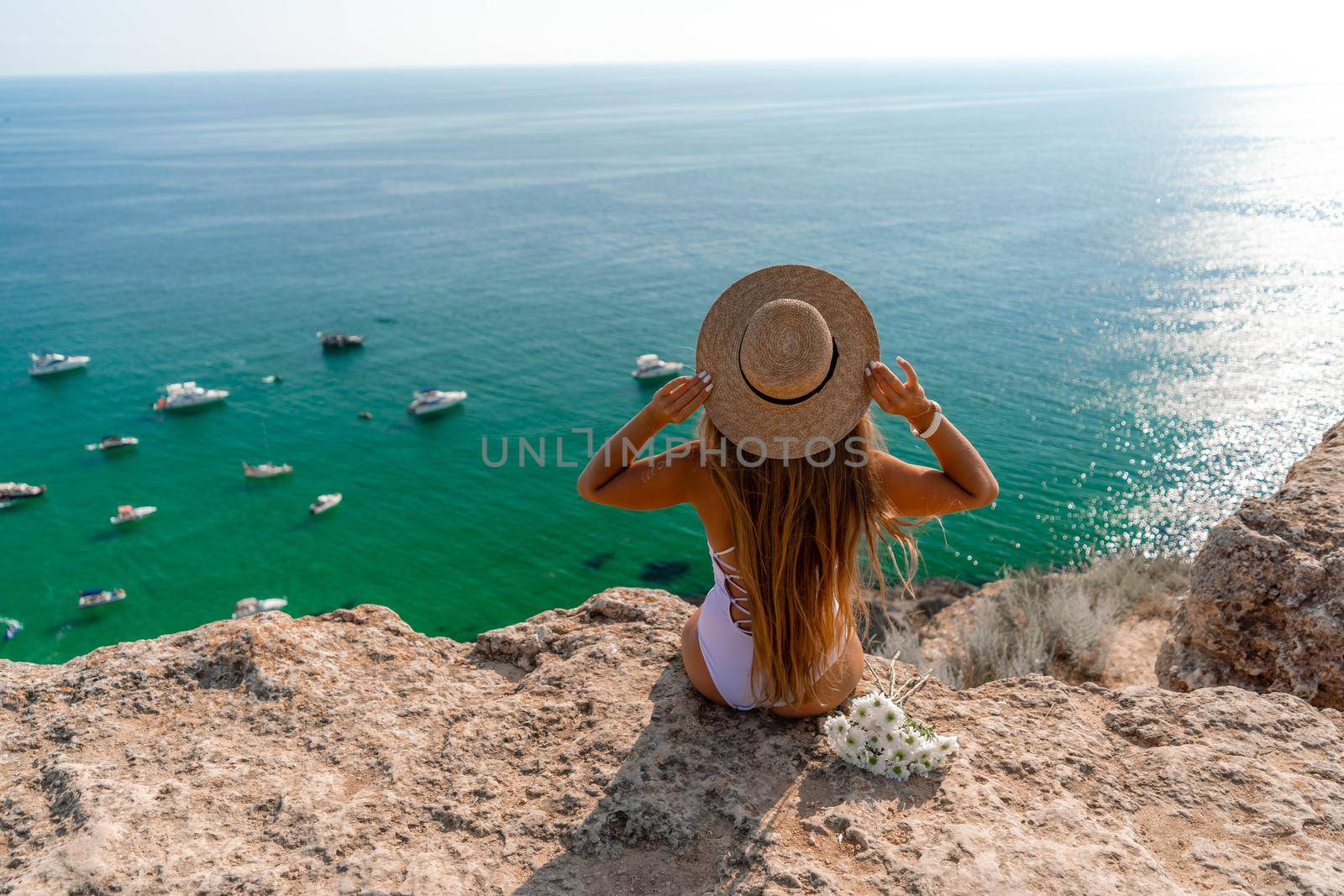 Beach holidays. A hot beautiful woman in a sun hat and a white bikini, sitting with her hands raised to her head, enjoying the view of the ocean on a hot summer day. by Matiunina