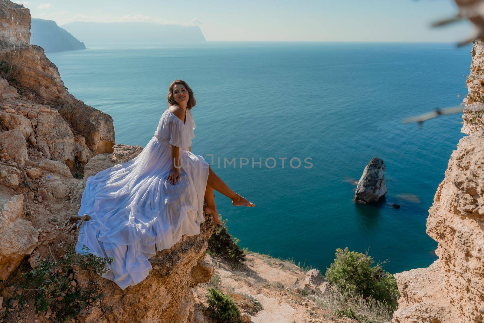 A beautiful young woman in a white light dress with long legs stands on the edge of a cliff above the sea waving a white long dress, against the background of the blue sky and the sea. by Matiunina