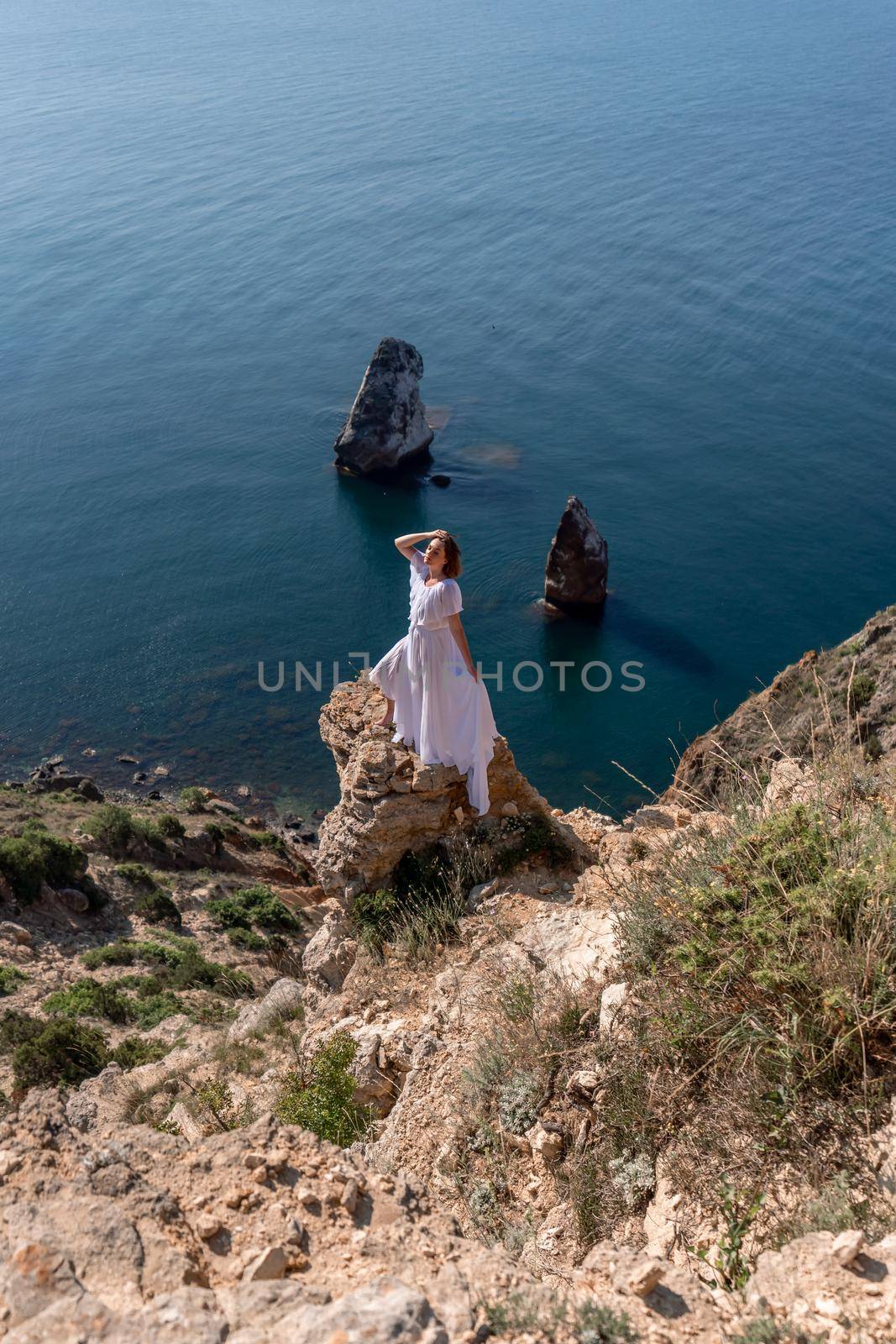 A beautiful young woman in a white light dress with long legs stands on the edge of a cliff above the sea waving a white long dress, against the background of the blue sky and the sea