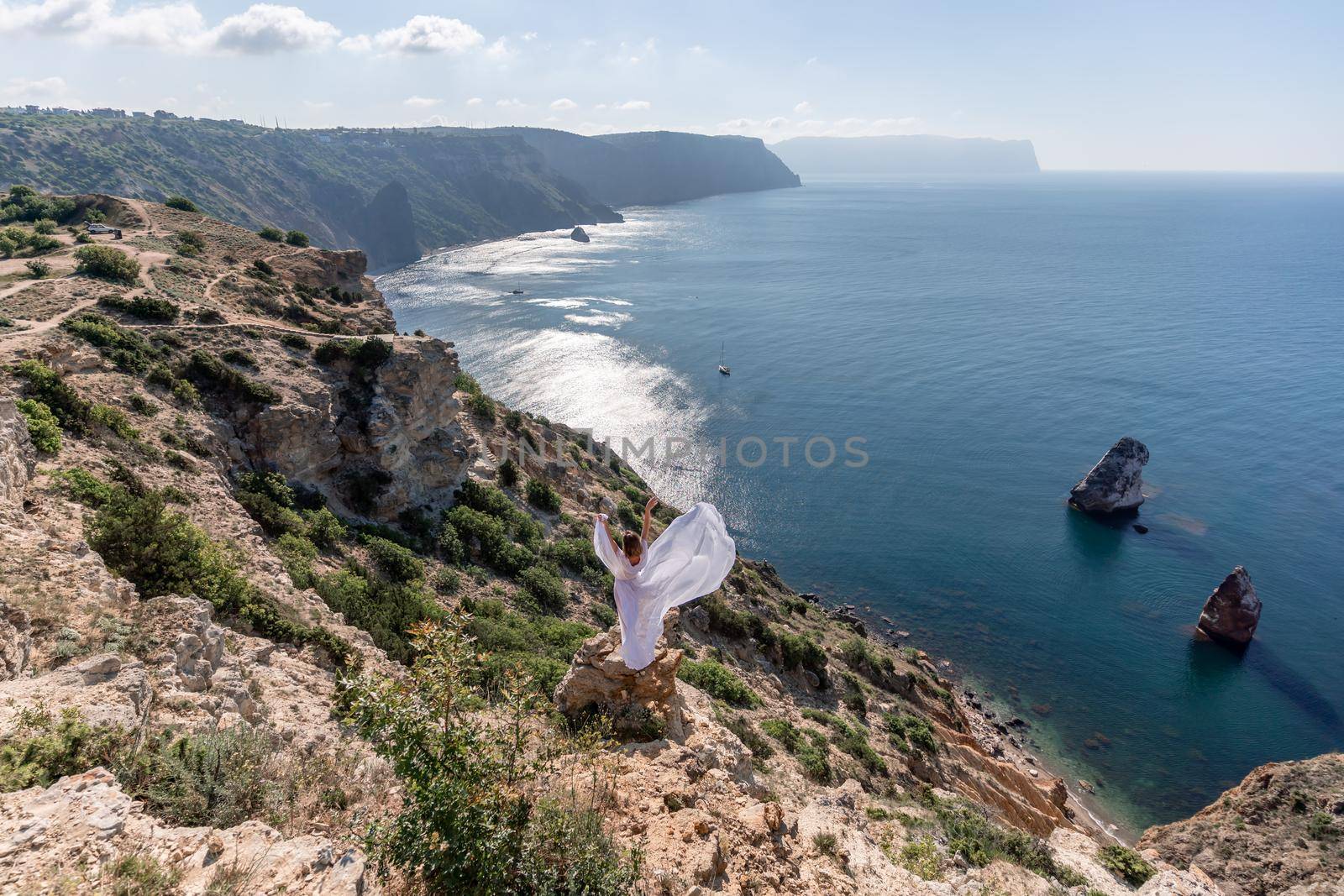 A beautiful young woman in a white light dress with long legs stands on the edge of a cliff above the sea waving a white long dress, against the background of the blue sky and the sea. by Matiunina