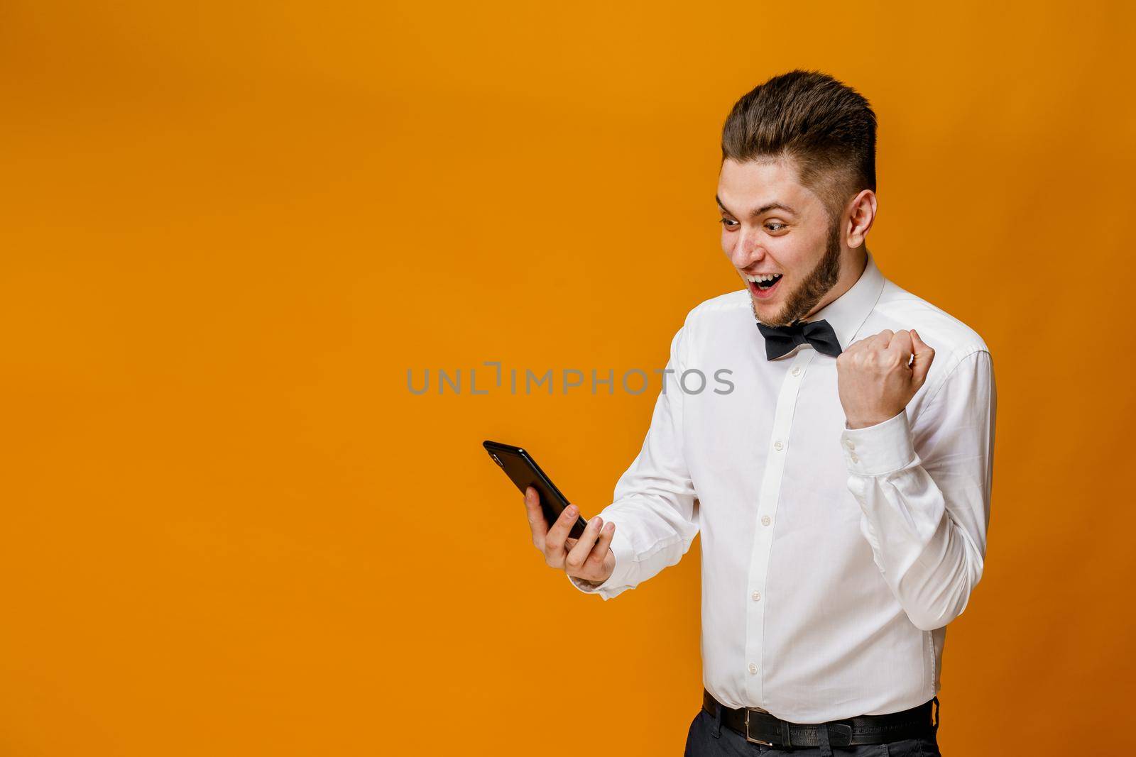Surprised handsome man wins on line bet. Caucasian man looks into the phone . Isolated orange backround. On-line work at home quarantine concept. by Rabizo