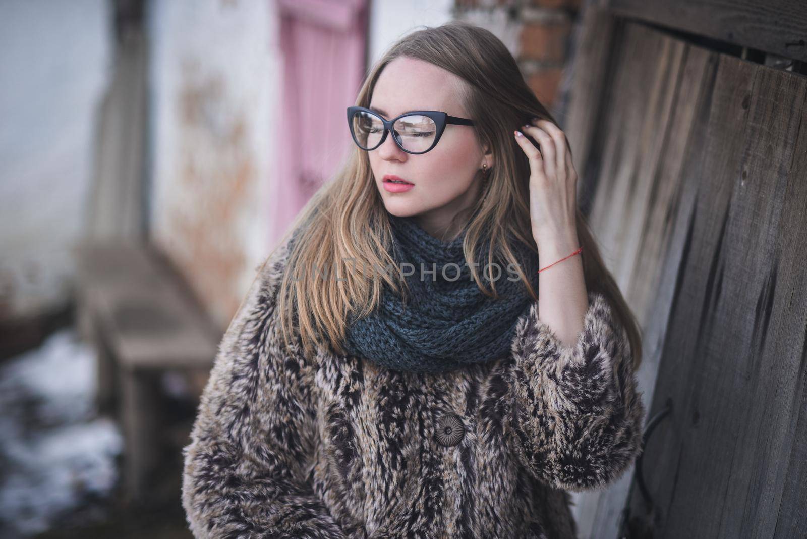 Girl blonde in cat eyes glasses in an artificial faux fur coat posing. Lying snow on the background. Red orange pink tones in the photo. Around the old shabby abandoned buildings. Cold winter