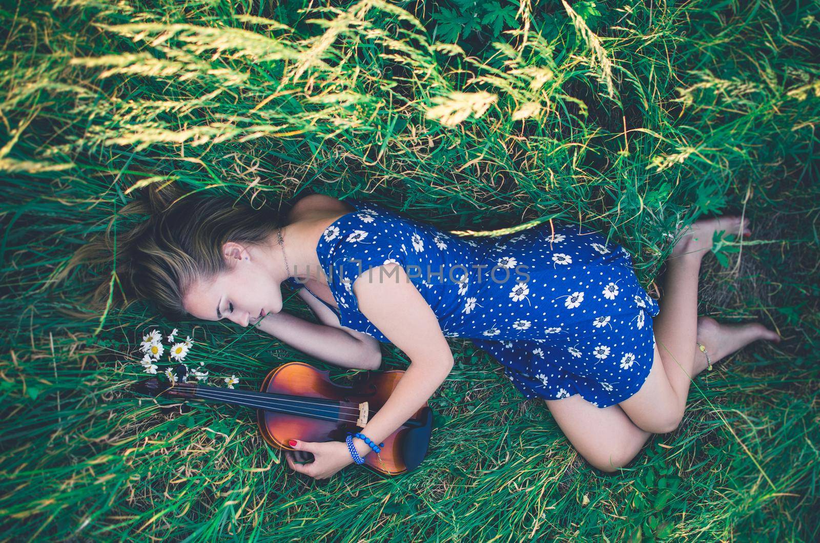 Young slim fair-skinned girl blonde beautiful lies in tall grass. Top view. Girl in a blue short tight dress with a print of daisies. A bouquet and lonely daisies scattered around.A woman hugs violin.